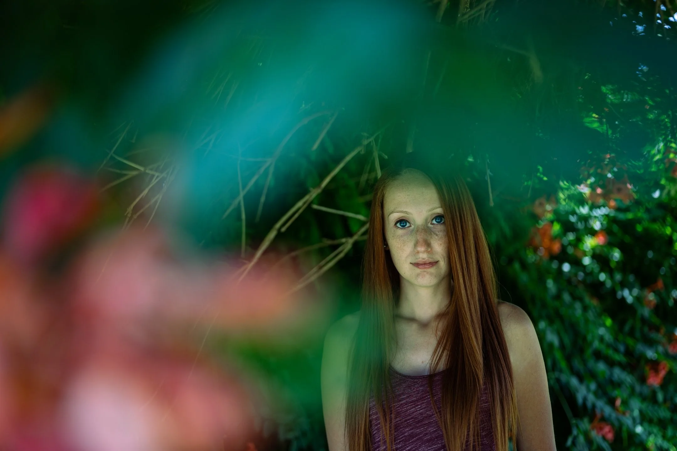 Young woman with red hair stands in a lush green setting, partially obscured by blurred foliage.