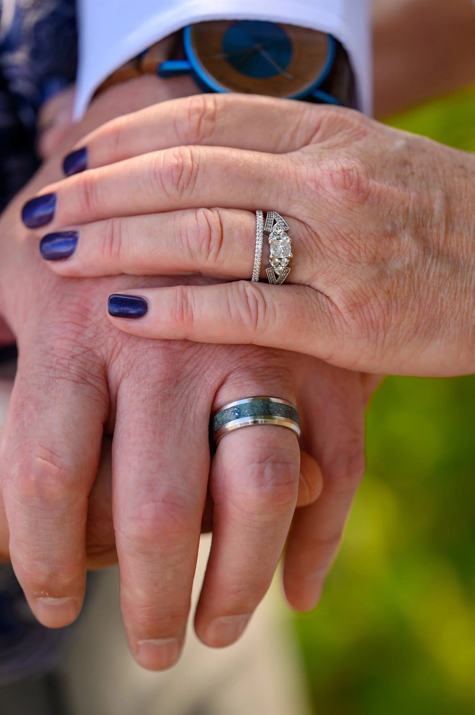 Close-up of a couple's hands with wedding rings, one with a diamond engagement ring and painted nails. A watch is partially visible.
