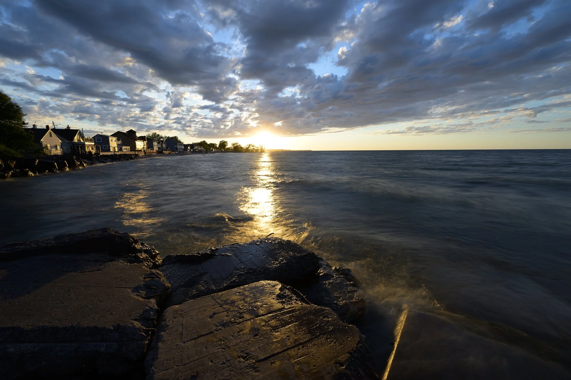 Sunset over a lake with rocky shore, houses, and cloudy sky.