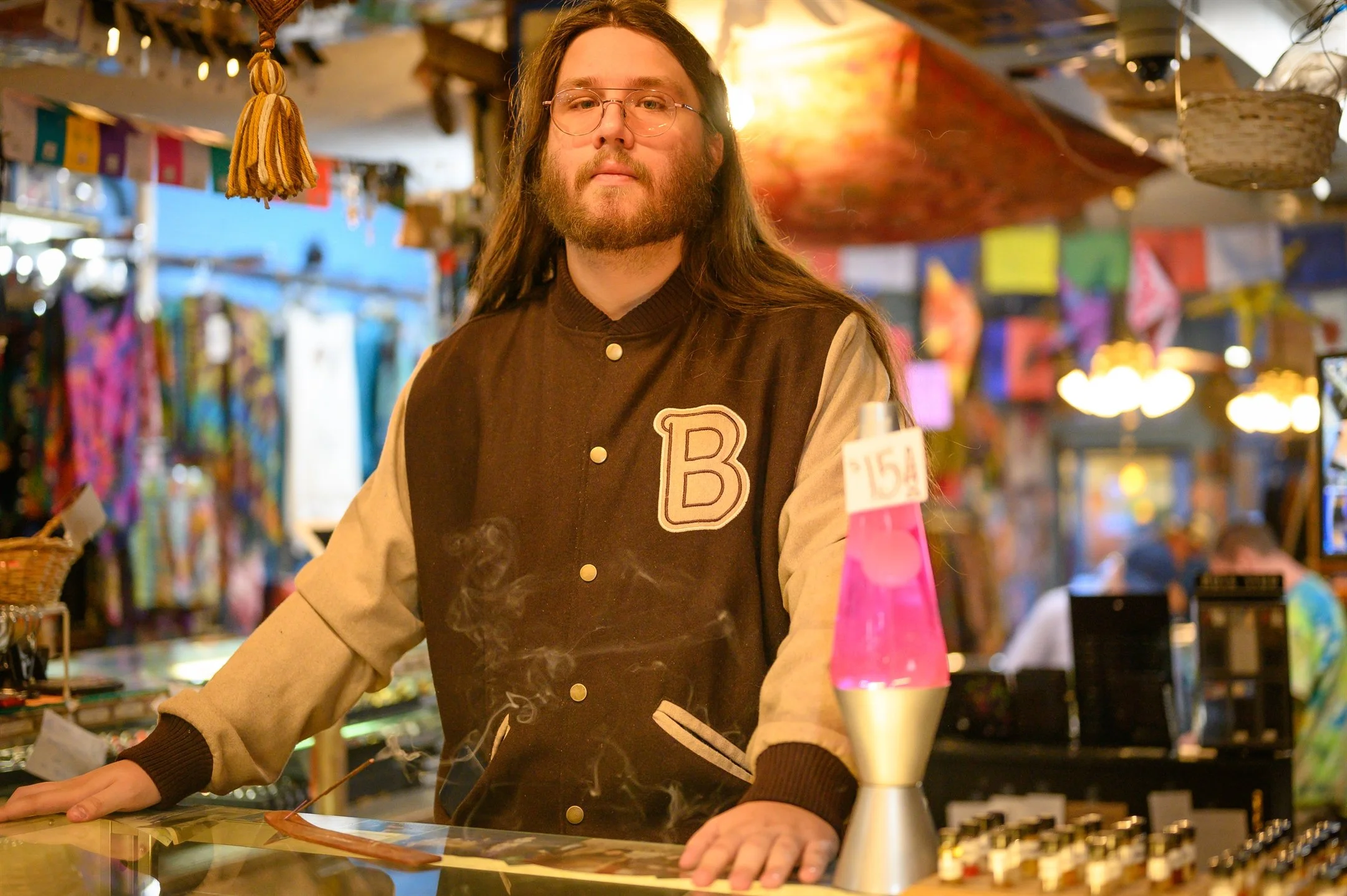 Person with long hair and glasses wearing a letterman jacket in a colorful store, standing by a pink lava lamp and incense.