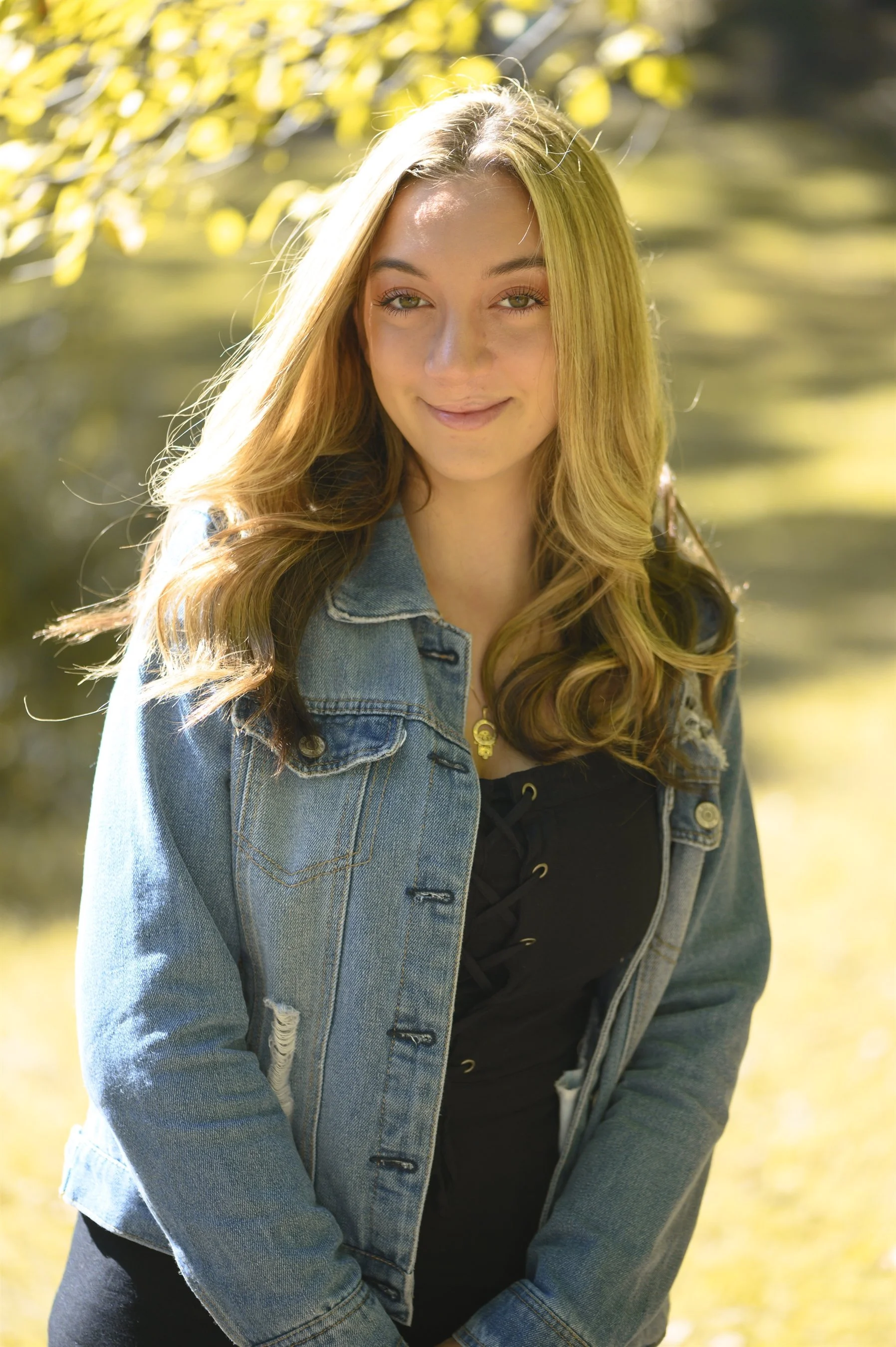 Young woman with long blonde hair wearing a denim jacket and black top, standing outdoors in soft natural light.