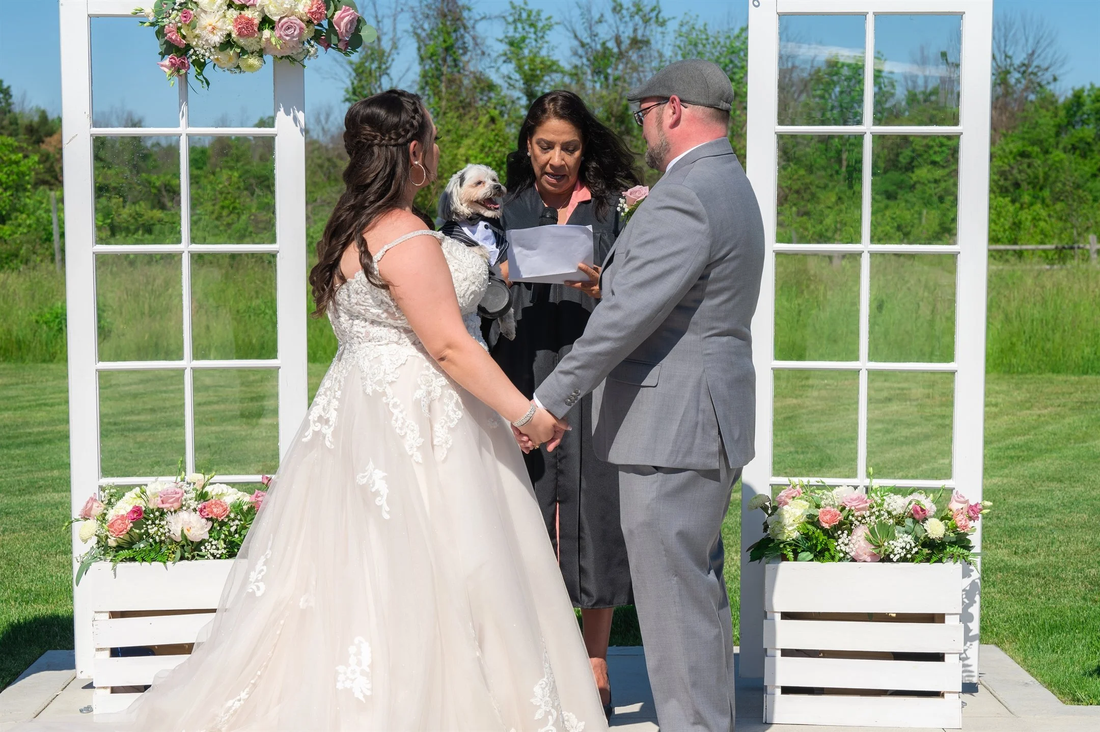 A wedding ceremony with a bride in a lace gown and a groom in a gray suit holding hands, standing in front of an officiant. The officiant is holding a small dog dressed in a tuxedo. The setting includes floral arrangements and a decorative arch.