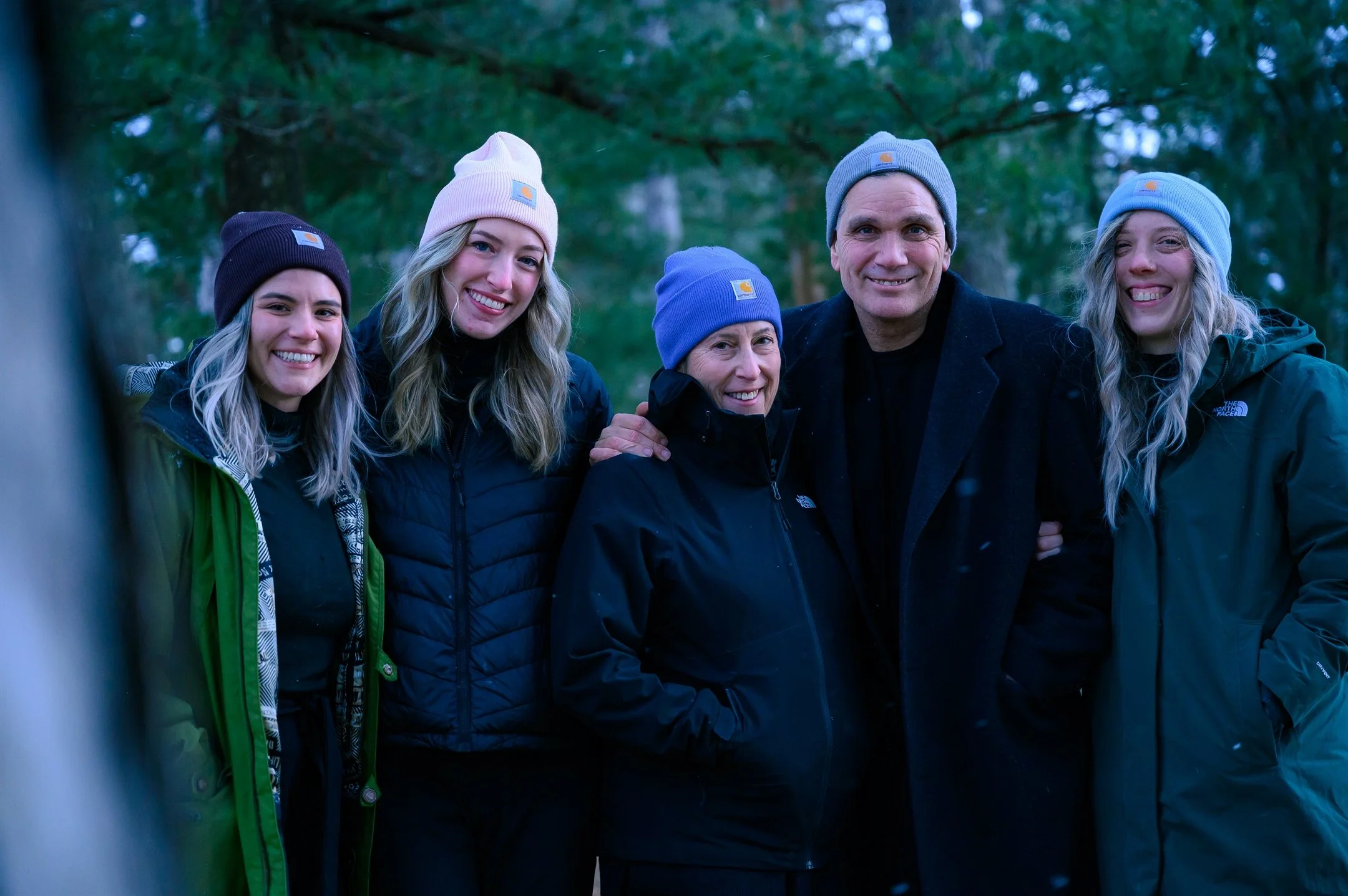 A group of five people outdoors, wearing winter clothing and beanies, smiling at the camera.