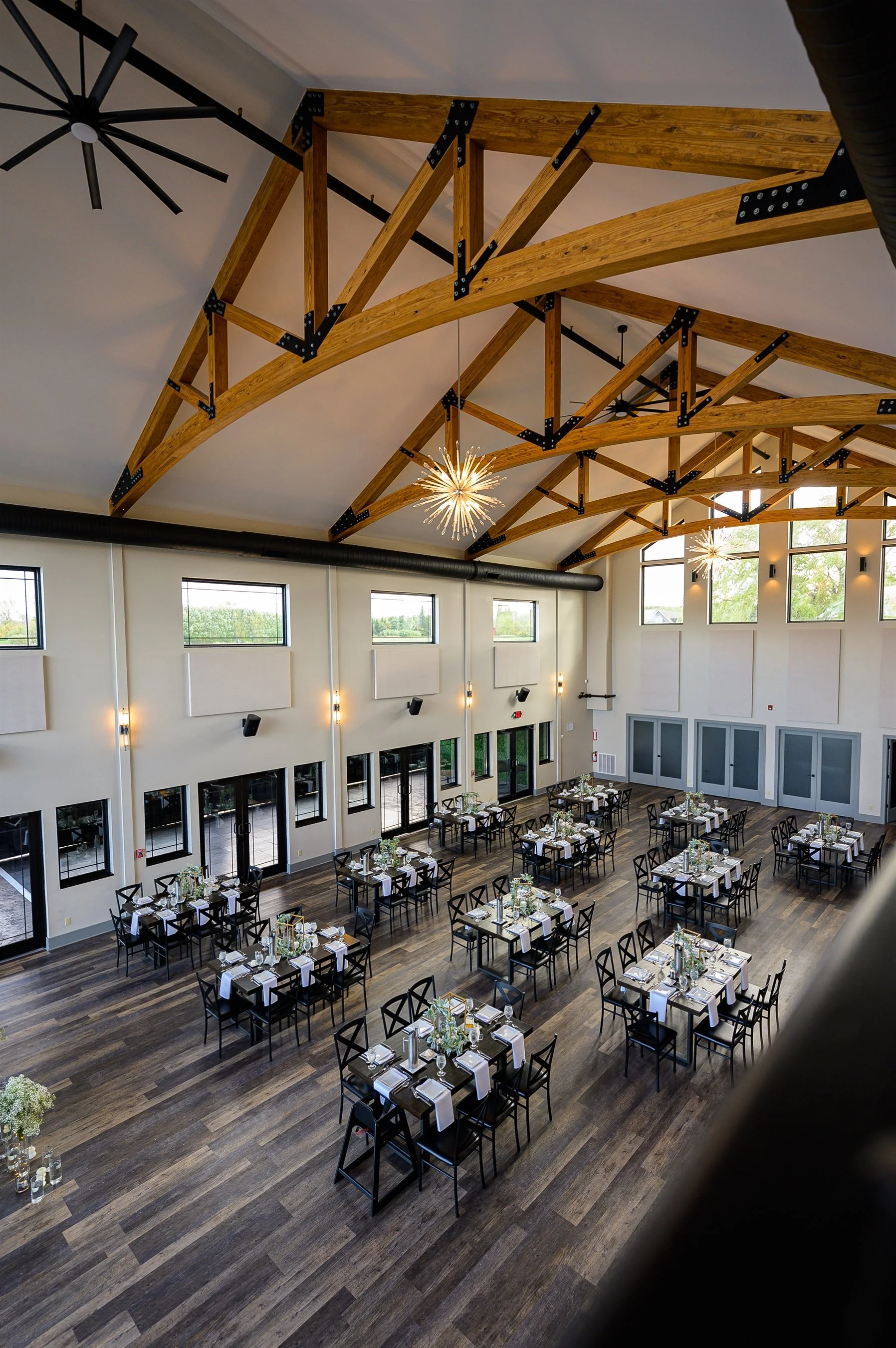 Elegant event venue interior with wooden ceiling beams, modern chandeliers, set dining tables, and large windows allowing natural light.