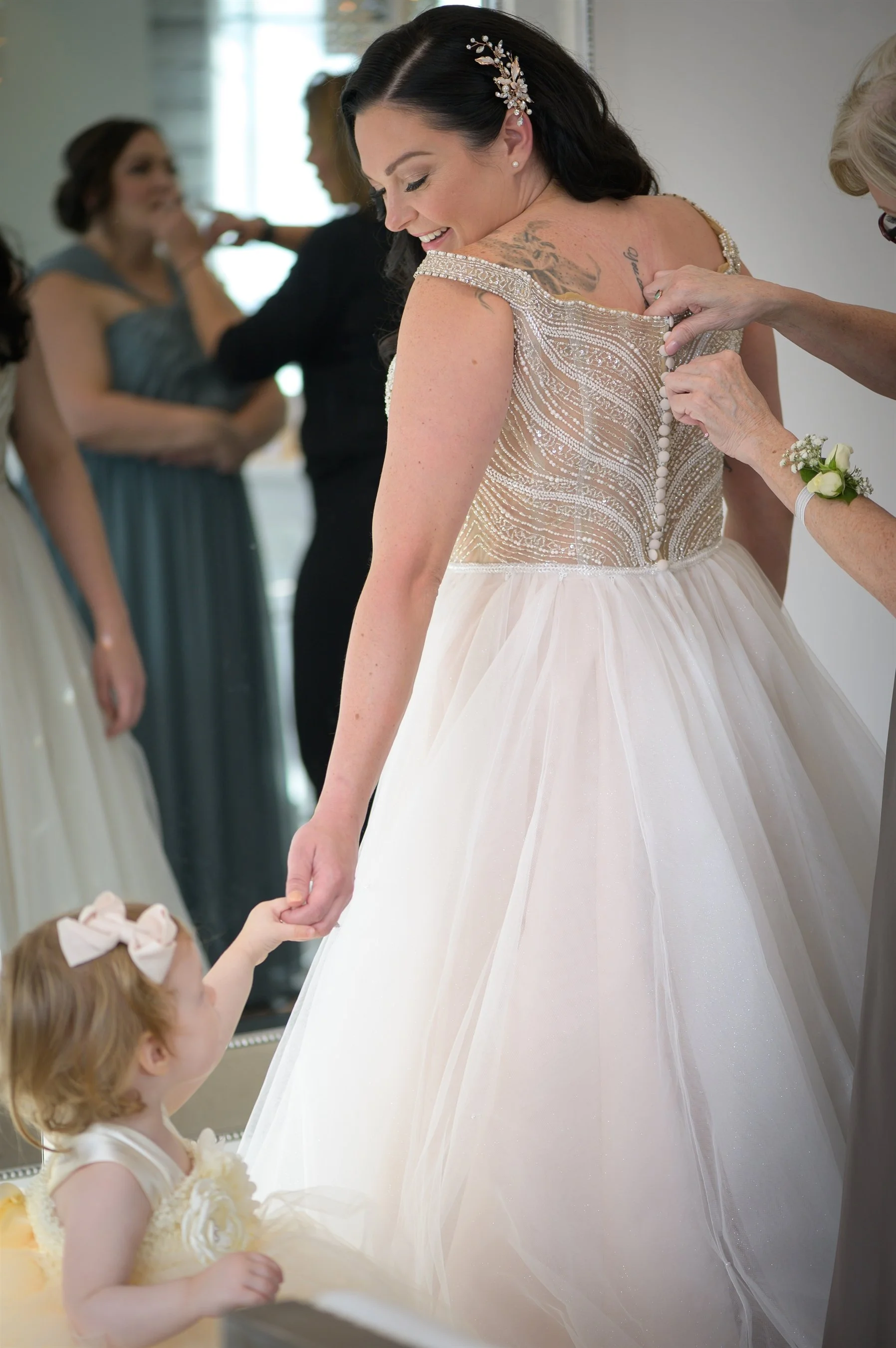 Bride in a white wedding dress being helped by an older woman, holding hands with a young girl in a dress.