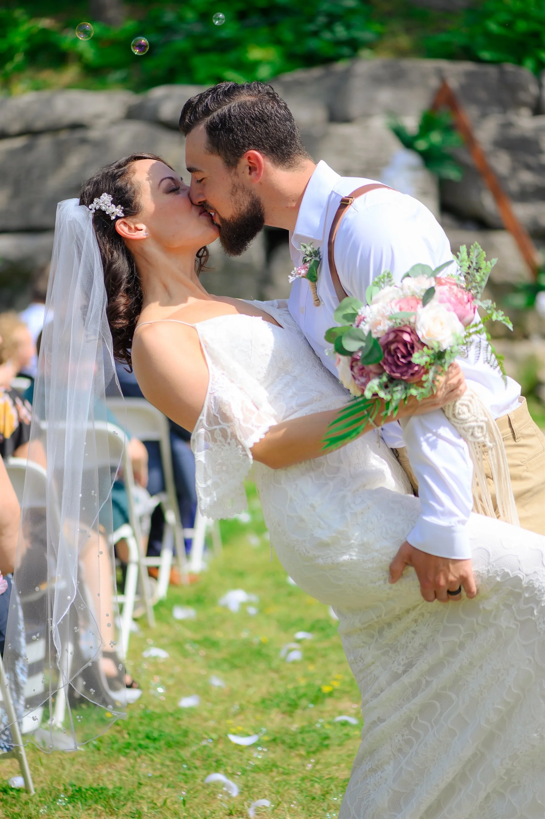 Bride and groom kissing outdoors at wedding ceremony, bride holding bouquet, groom dipping bride, both in formal attire.