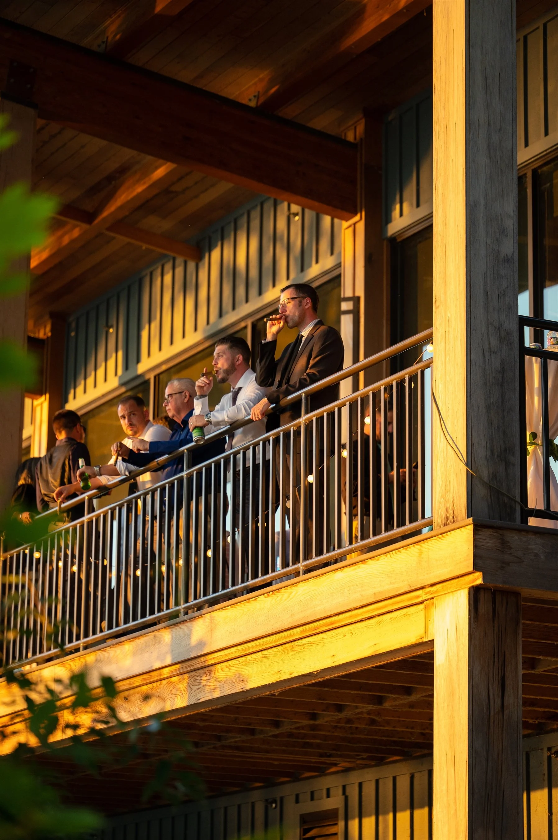Group of men on a wooden balcony, some holding drinks, during sunset.