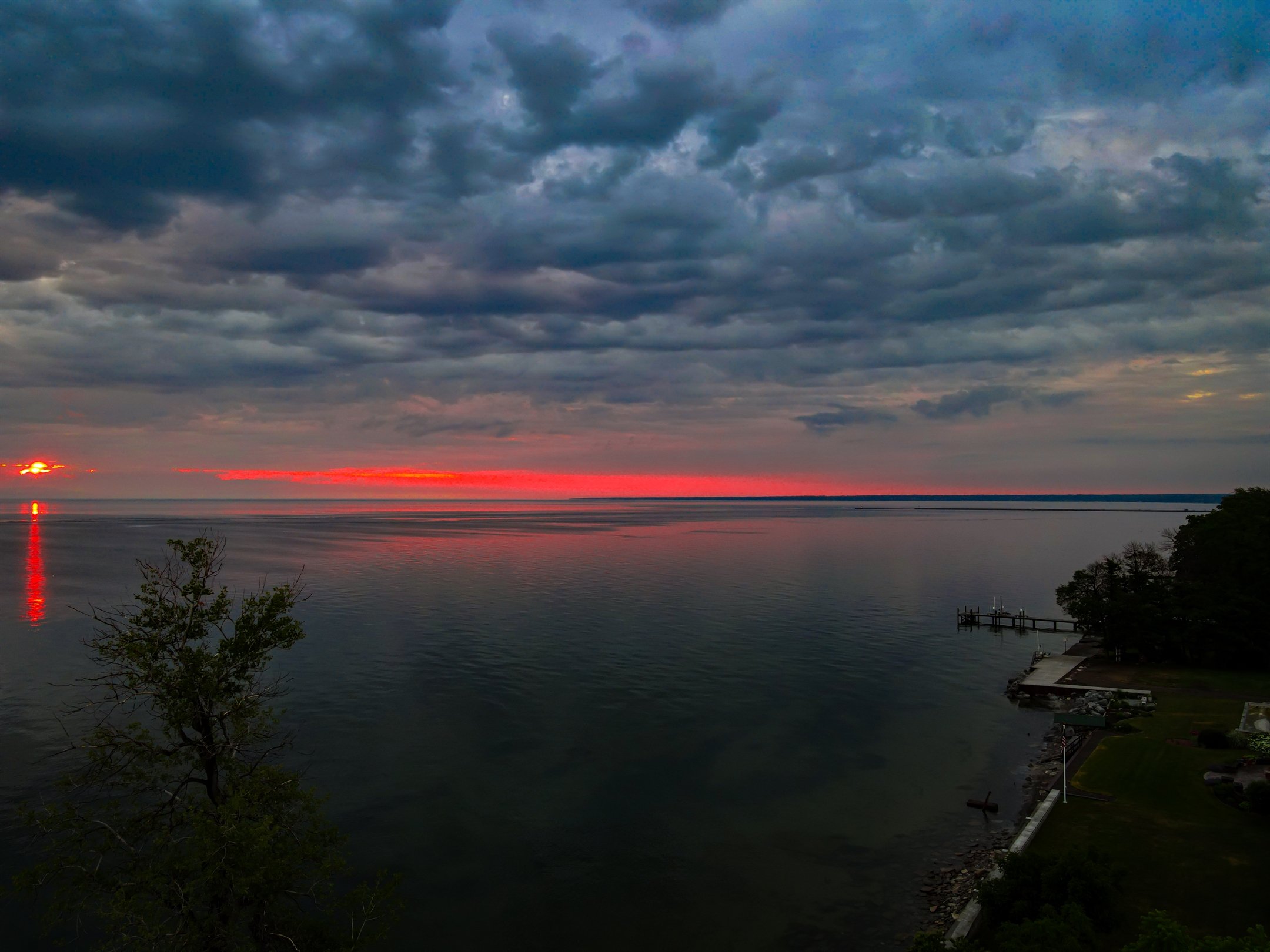Sunset over a lake with a tree in the foreground, dark clouds above, and orange-red reflection on the water.