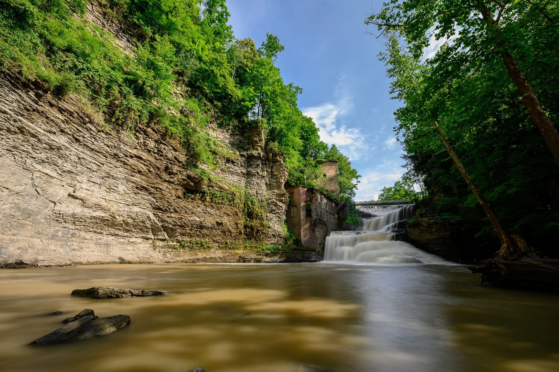 Scenic view of a waterfall cascading over rocky terrain surrounded by lush greenery and trees under a blue sky.