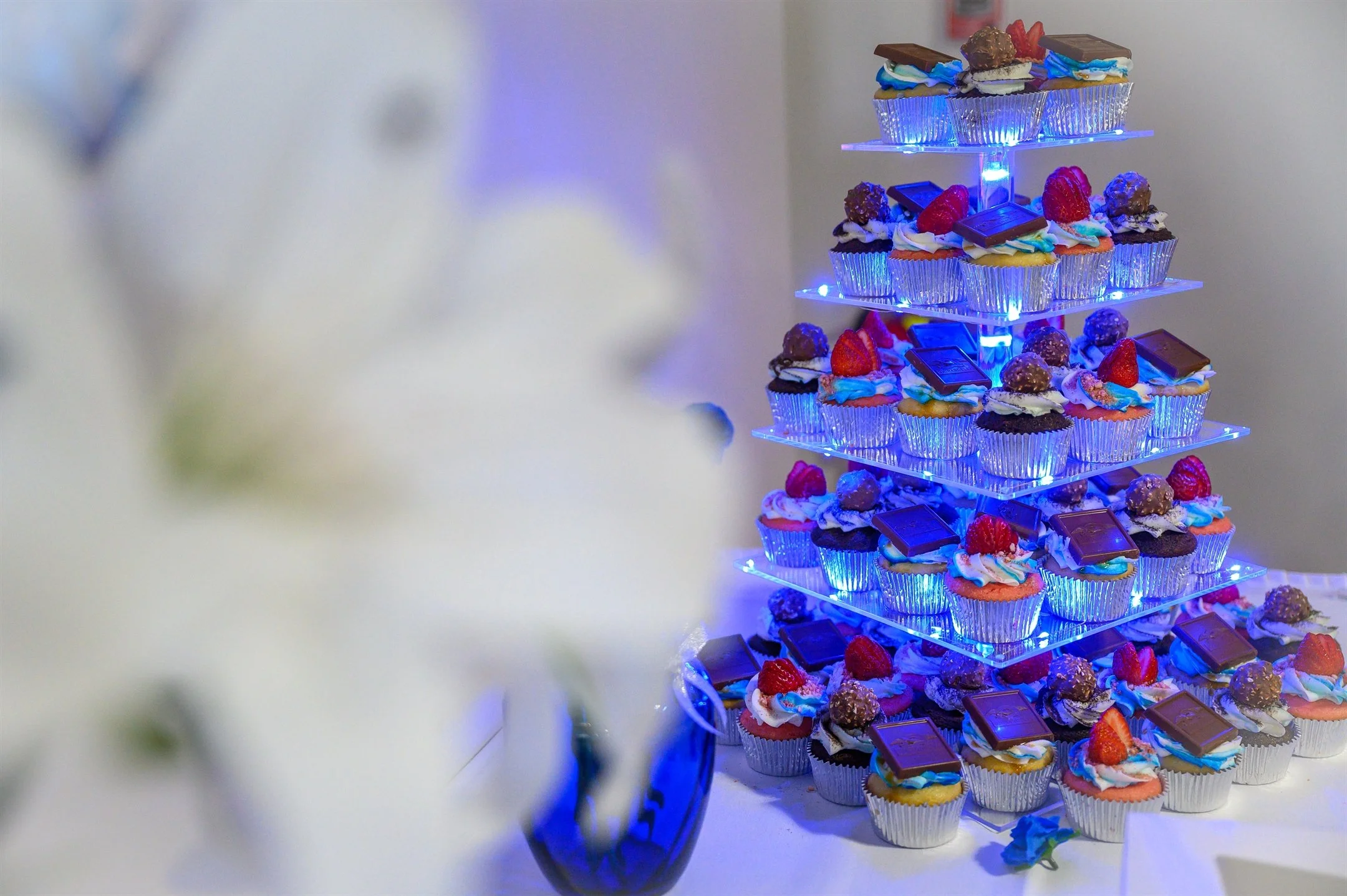 Tiered cupcake stand with decorated desserts, including chocolates and berries, illuminated with blue lights. Blurred white flowers in foreground.