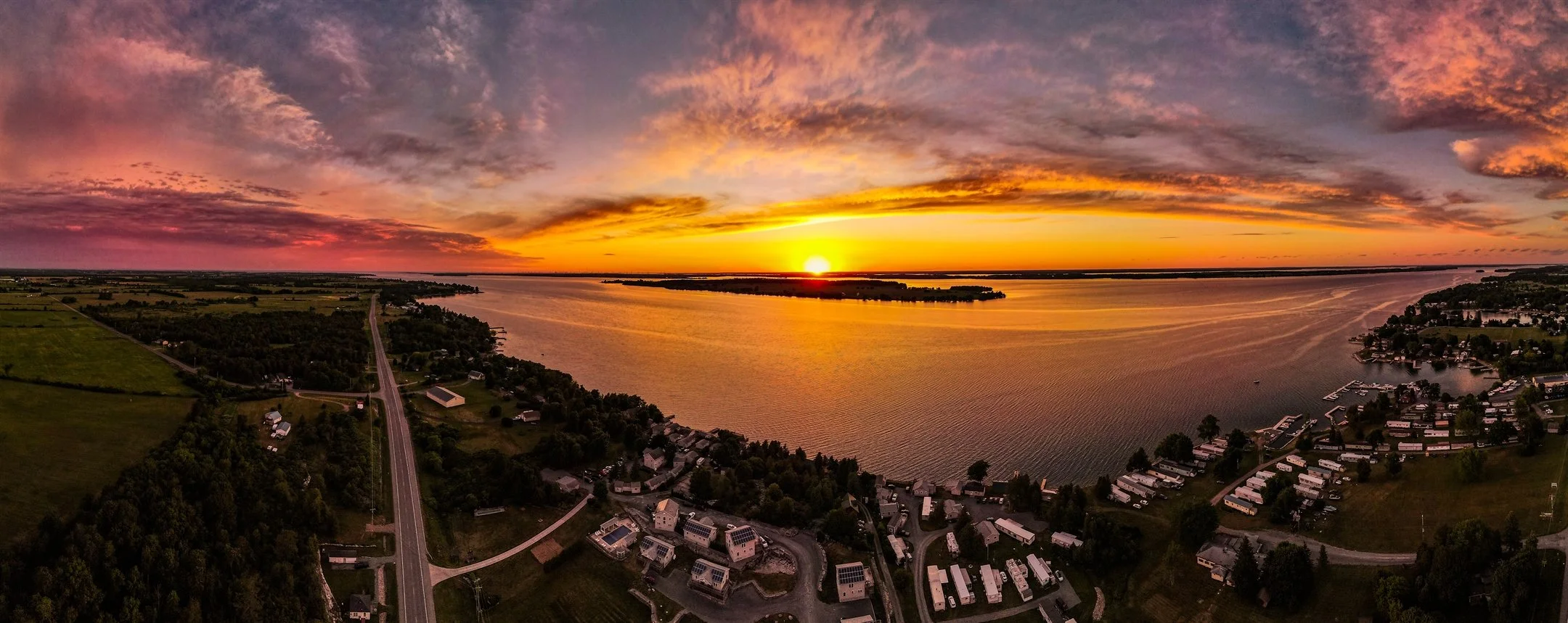Panoramic view of a colorful sunset over a large lake with nearby residential area and lush greenery.
