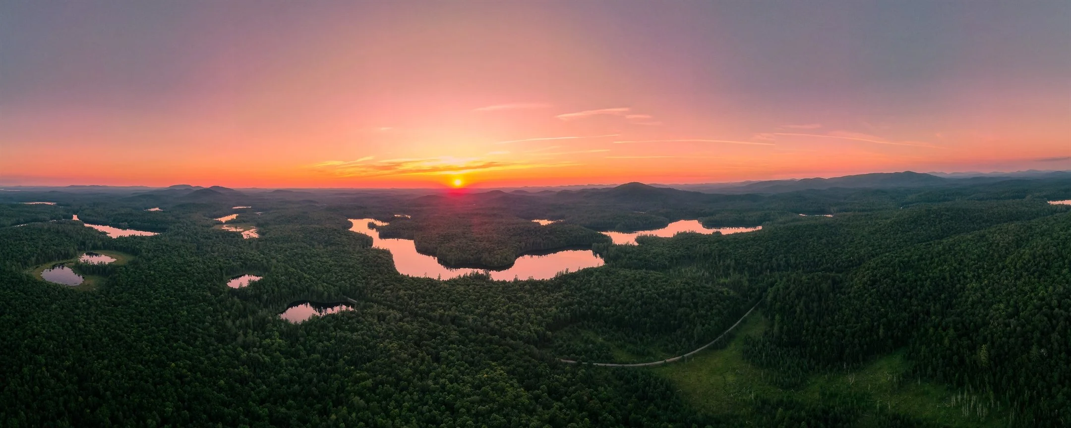 Aerial view of a landscape at sunset with forested areas and numerous lakes reflecting the colorful sky.