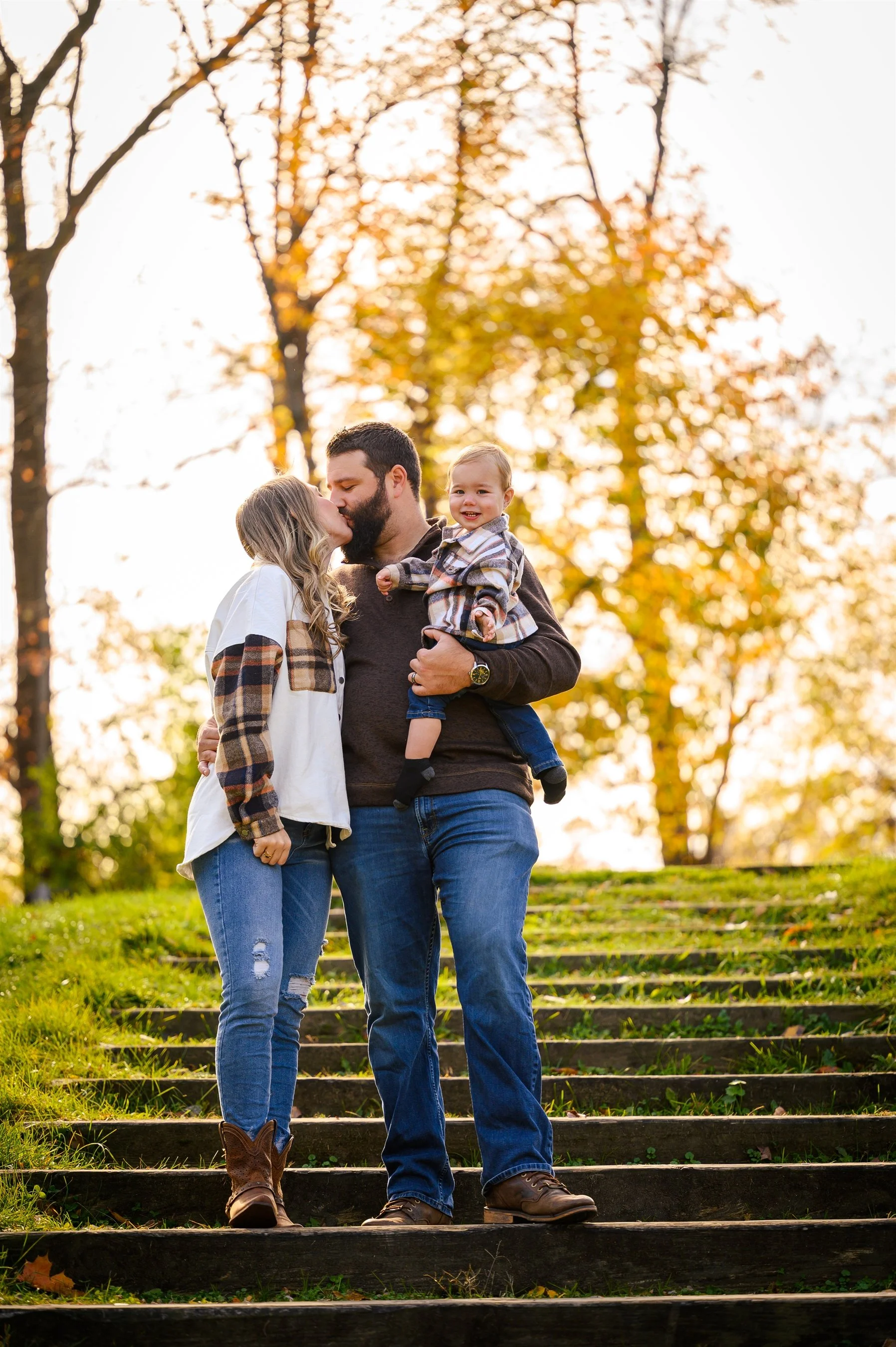 A couple kissing while holding a child, standing on outdoor steps with autumn trees in the background.
