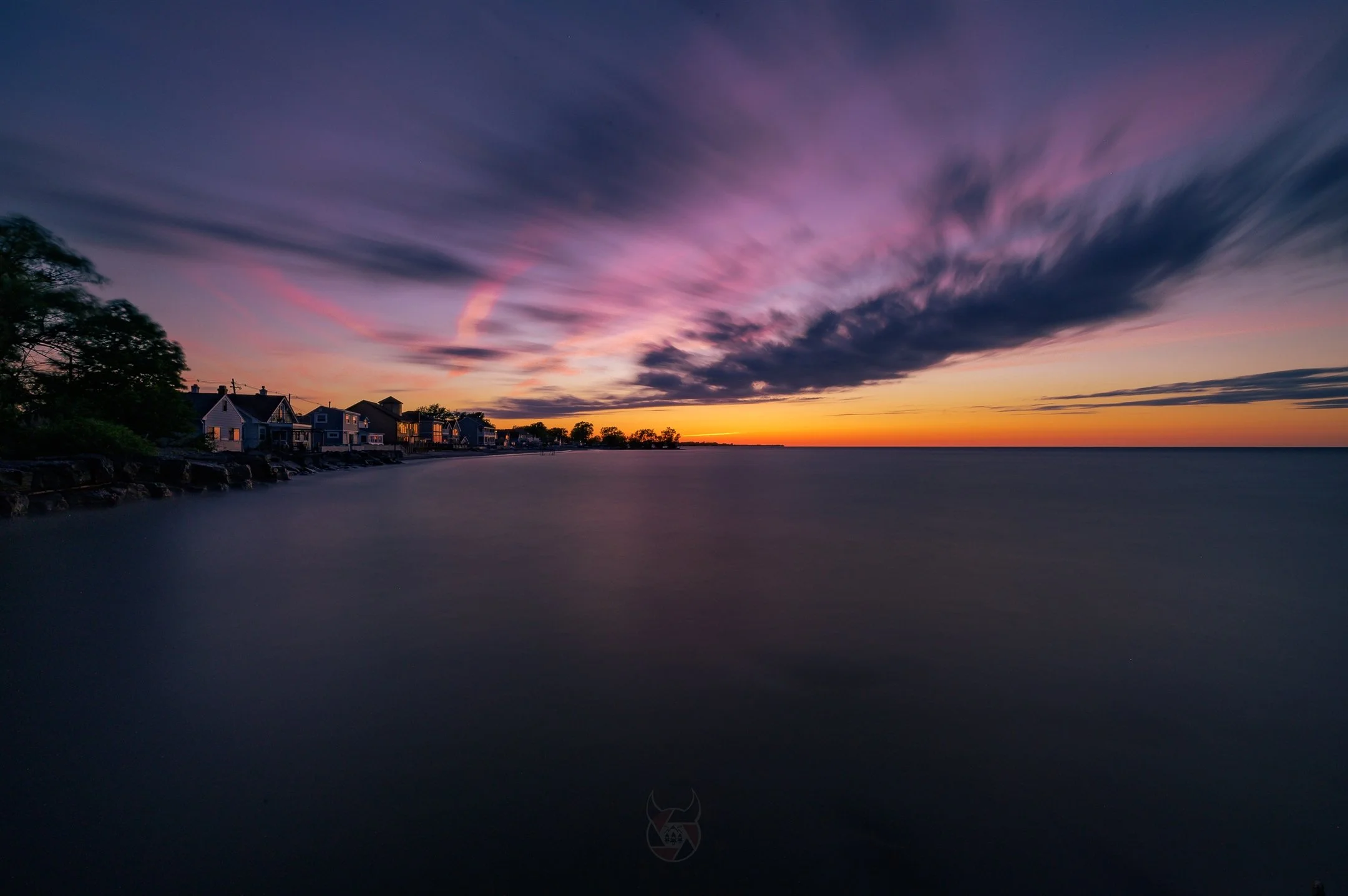 Long exposure of a picturesque sunset over a calm lake. Silhouetted shoreline with houses and trees on the left. Vibrant sky with shades of orange, purple, and blue, reflecting softly on the water.