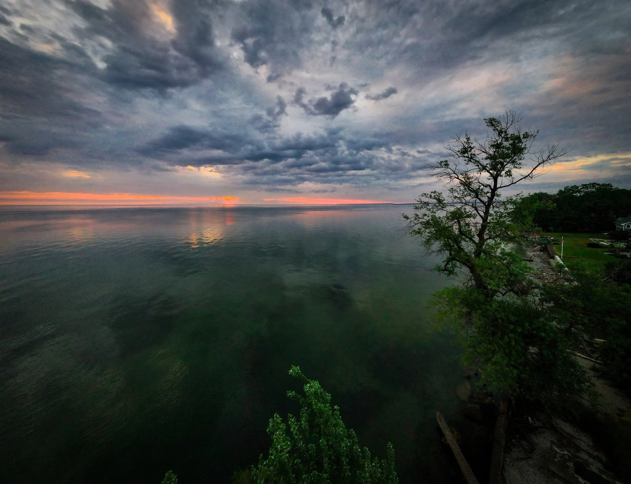 A scenic view of a calm lake with a partly cloudy sky during sunset, orange and pink hues reflecting on the water, and trees along the shoreline.