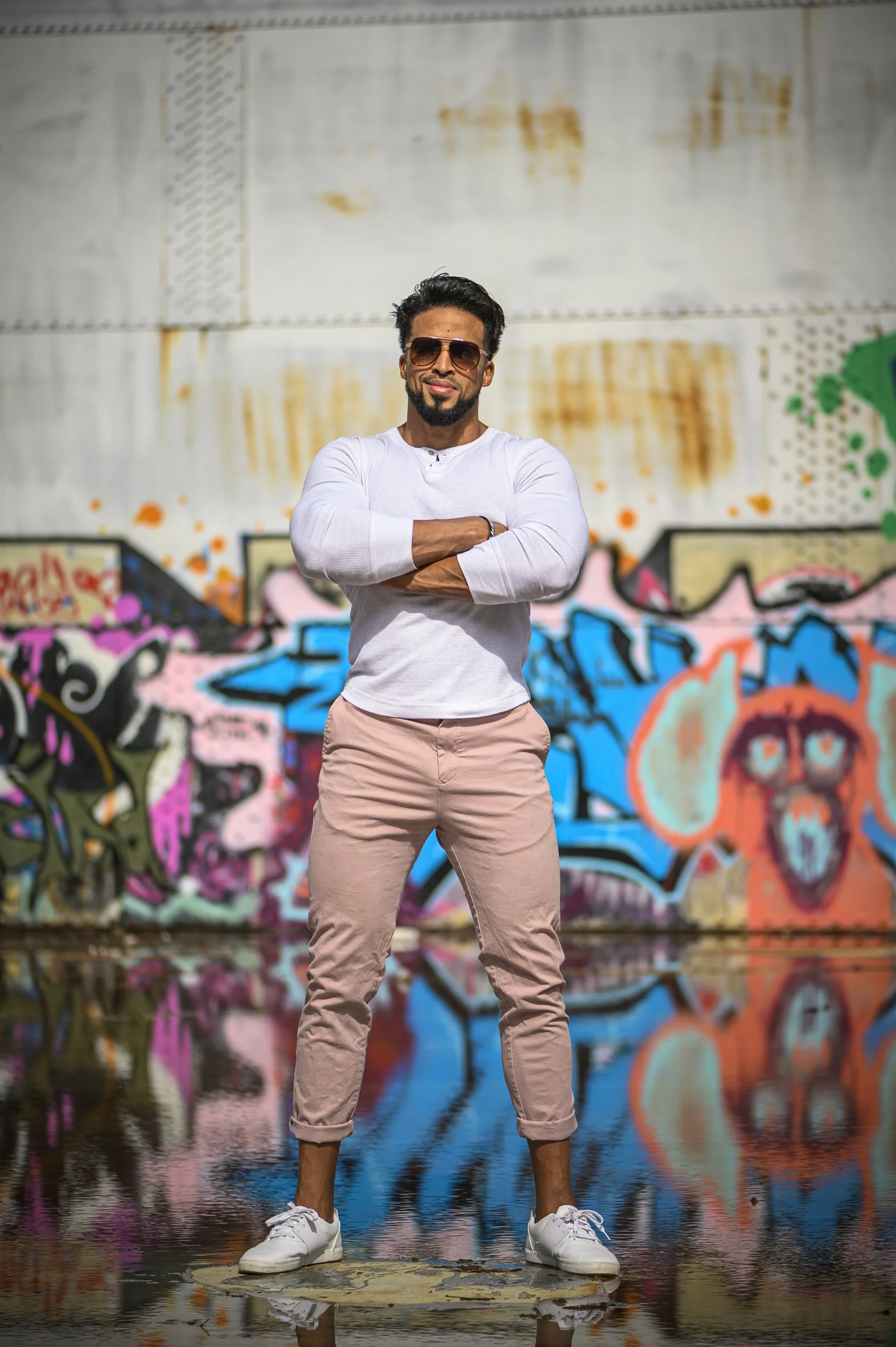 Man in white long-sleeve shirt and pink pants standing in front of colorful graffiti wall, with arms crossed and wearing sunglasses.