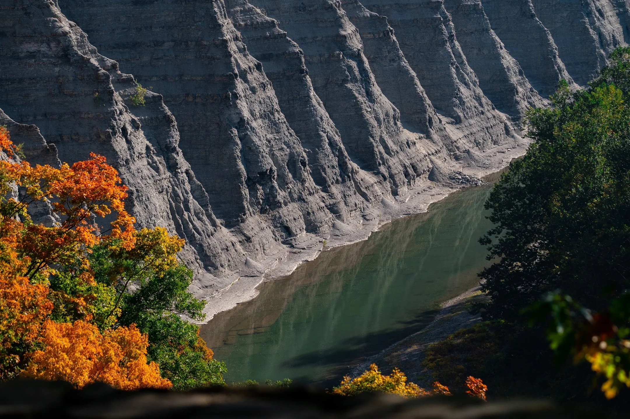 Steep rock cliffs over a calm river with autumn trees in the foreground.