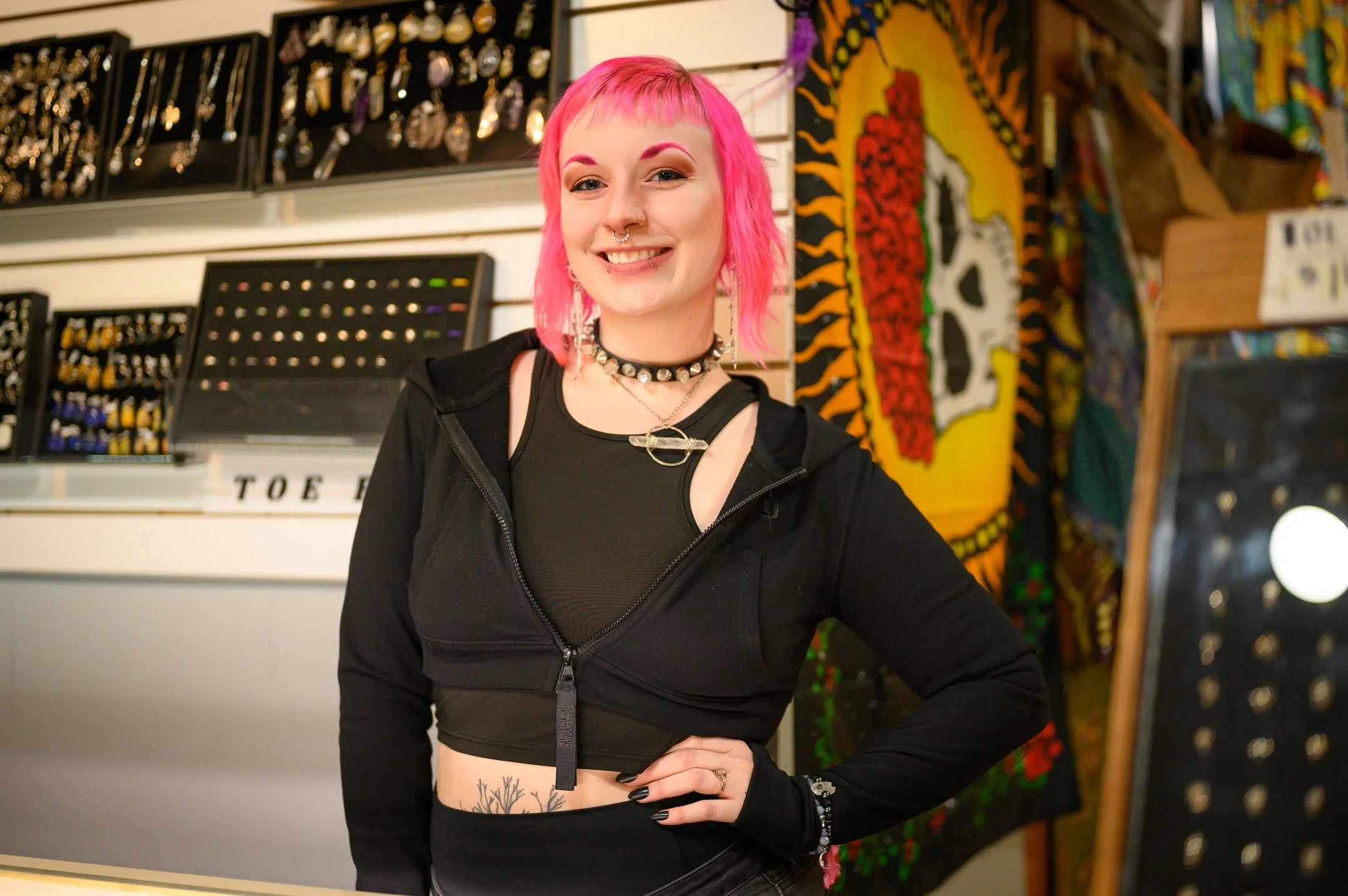 Person with pink hair, black crop top, and jewelry standing in front of store display with various accessories.