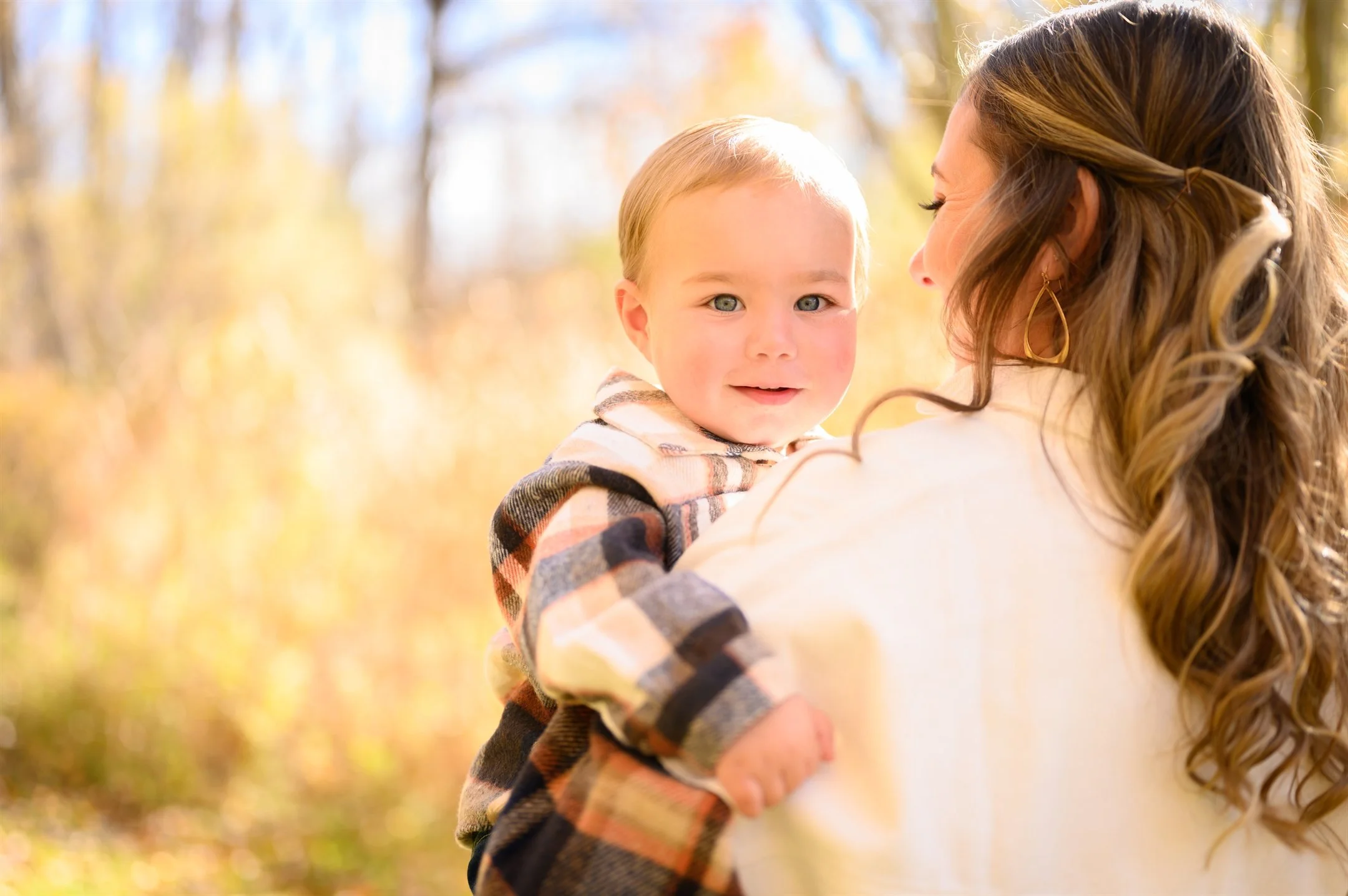 A woman holding a toddler outdoors in autumn with blurred fall foliage in the background.
