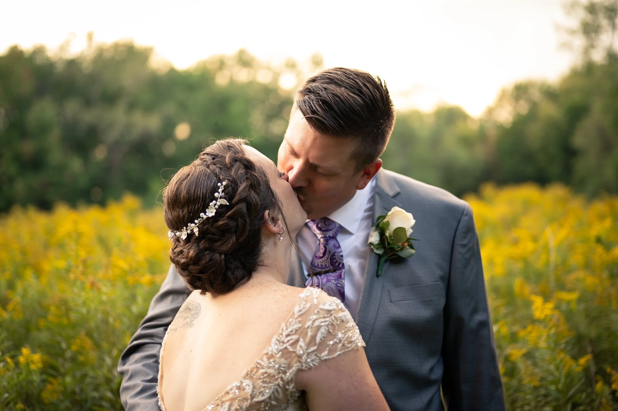 Bride in a lace dress and groom in a suit, kissing in a field with yellow flowers and trees in the background.