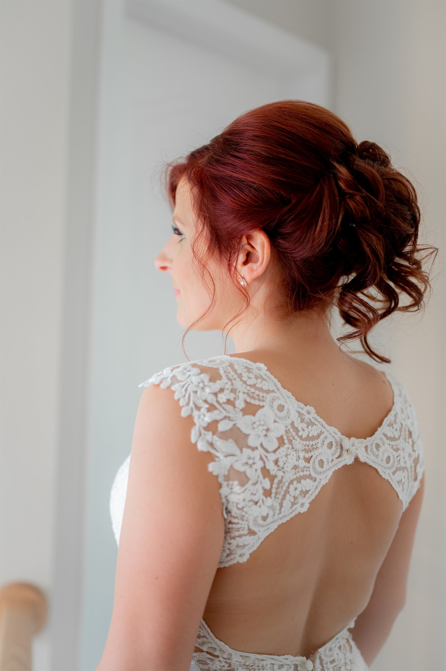 Bride facing away, wearing a lace wedding dress with open back and styled red hair.
