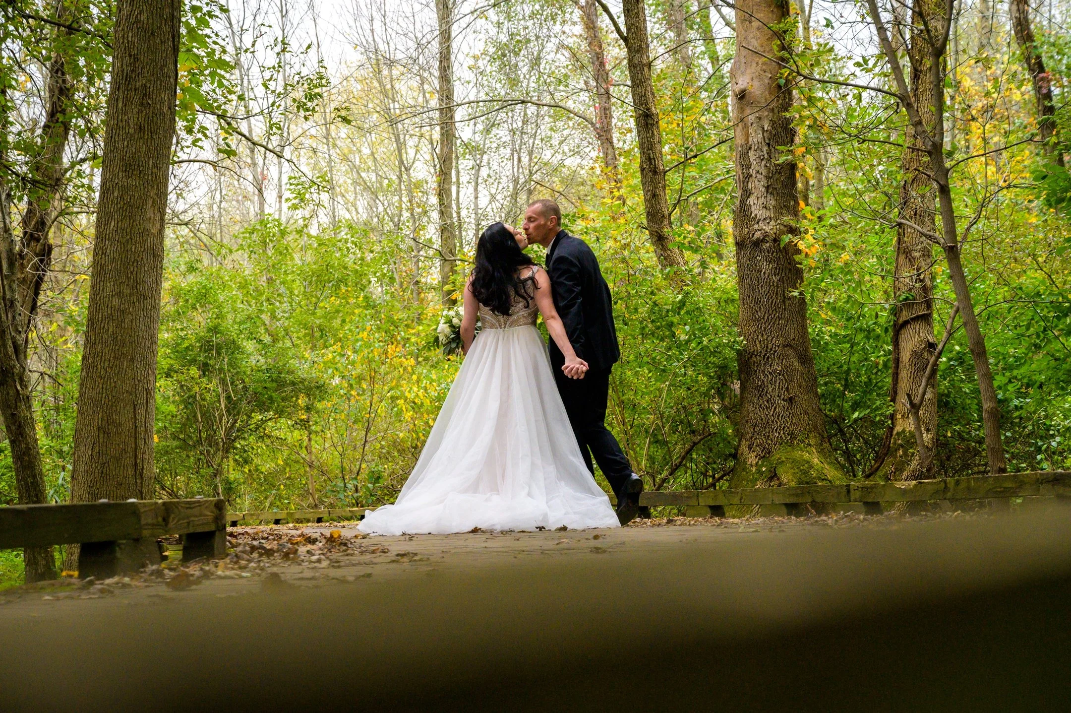 Bride and groom kissing on a forest path surrounded by trees and greenery.