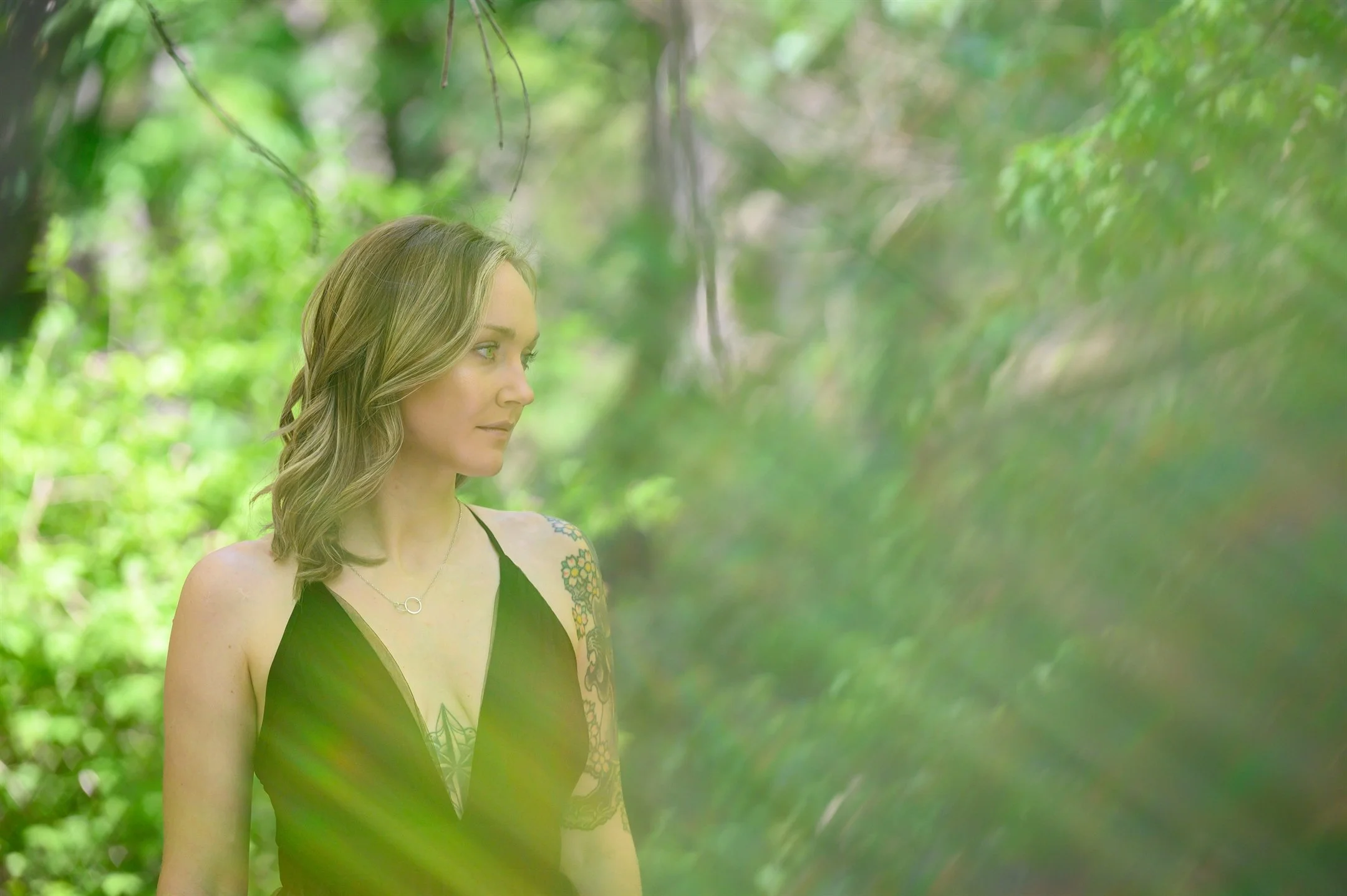 Woman with tattoos in a forest wearing a black dress, surrounded by green foliage.