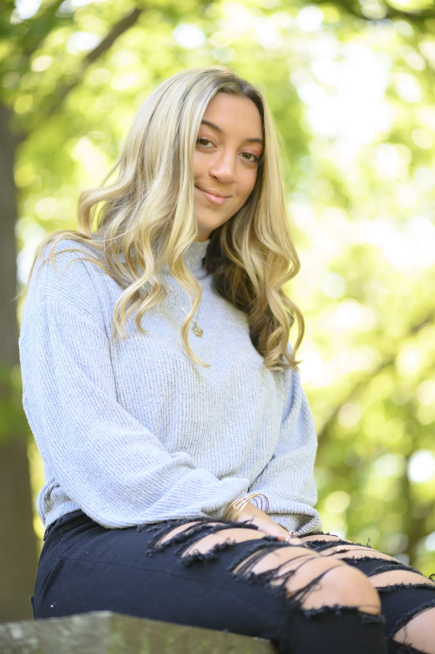 Woman with long blonde hair sitting outdoors, wearing a light gray sweater and ripped black jeans.