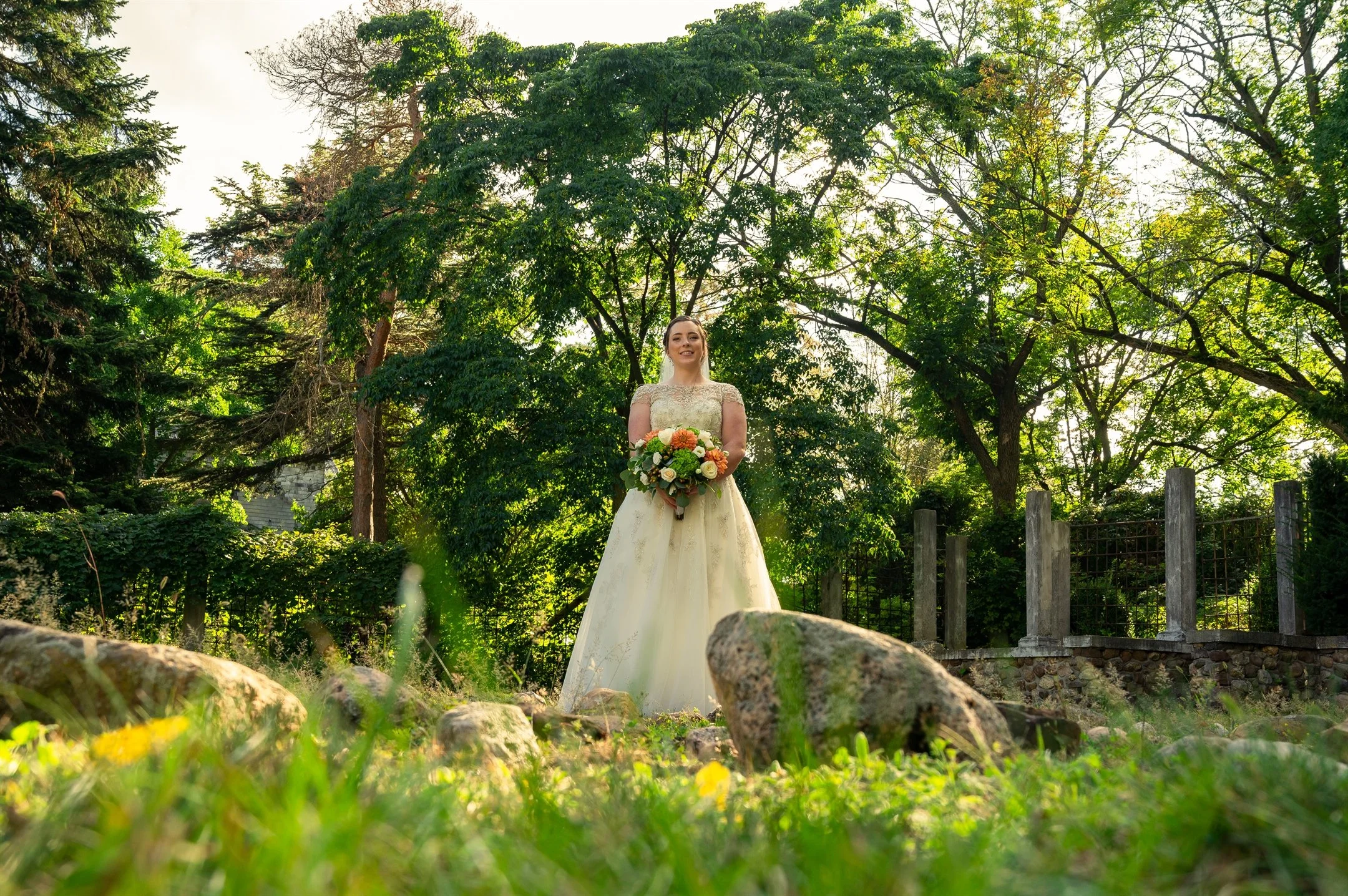 Bride holding a bouquet, standing in a garden with trees and stone pillars in the background.