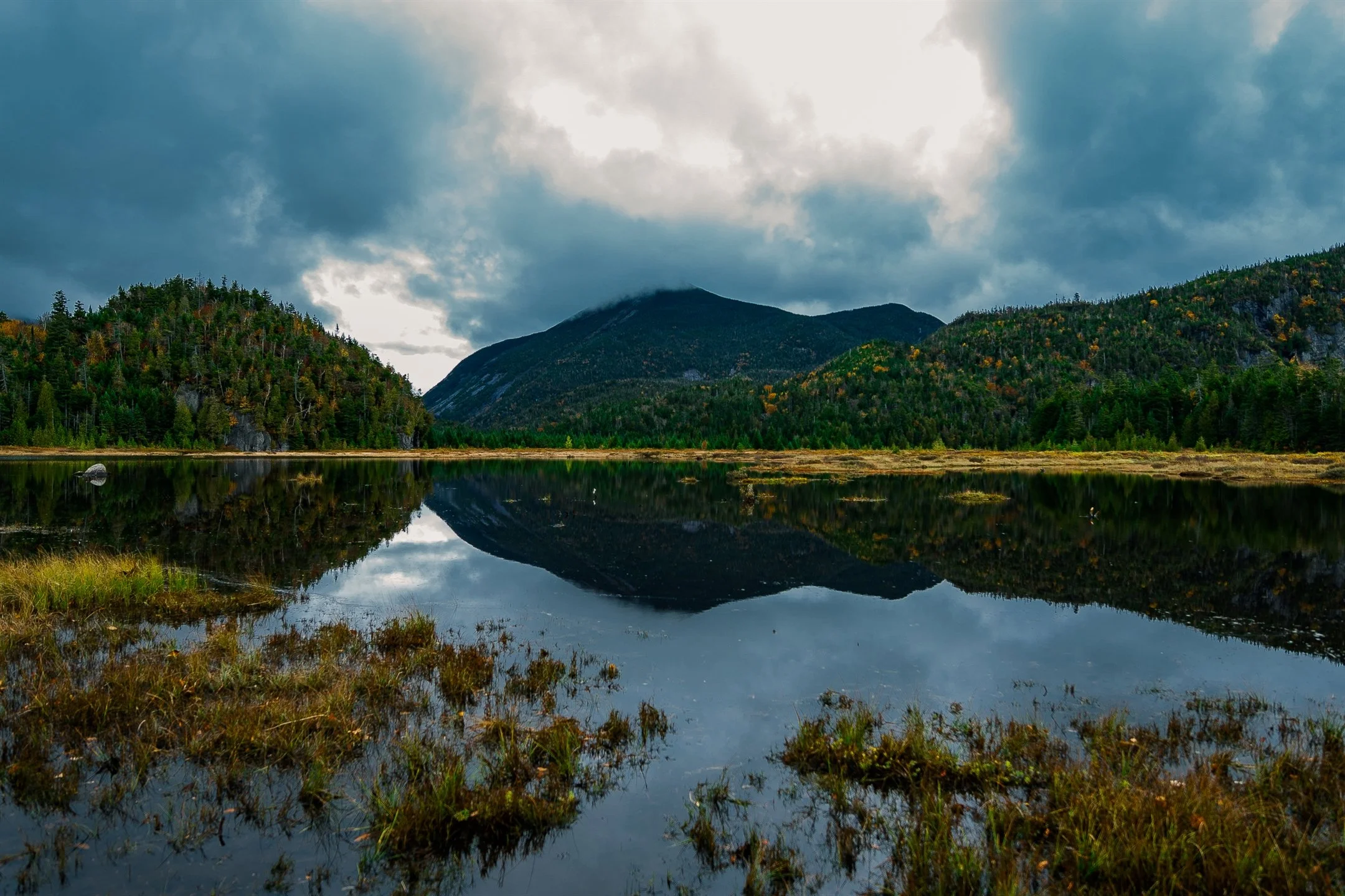 Scenic view of a calm lake reflecting surrounding mountains and cloudy sky, with lush forest on the shoreline.