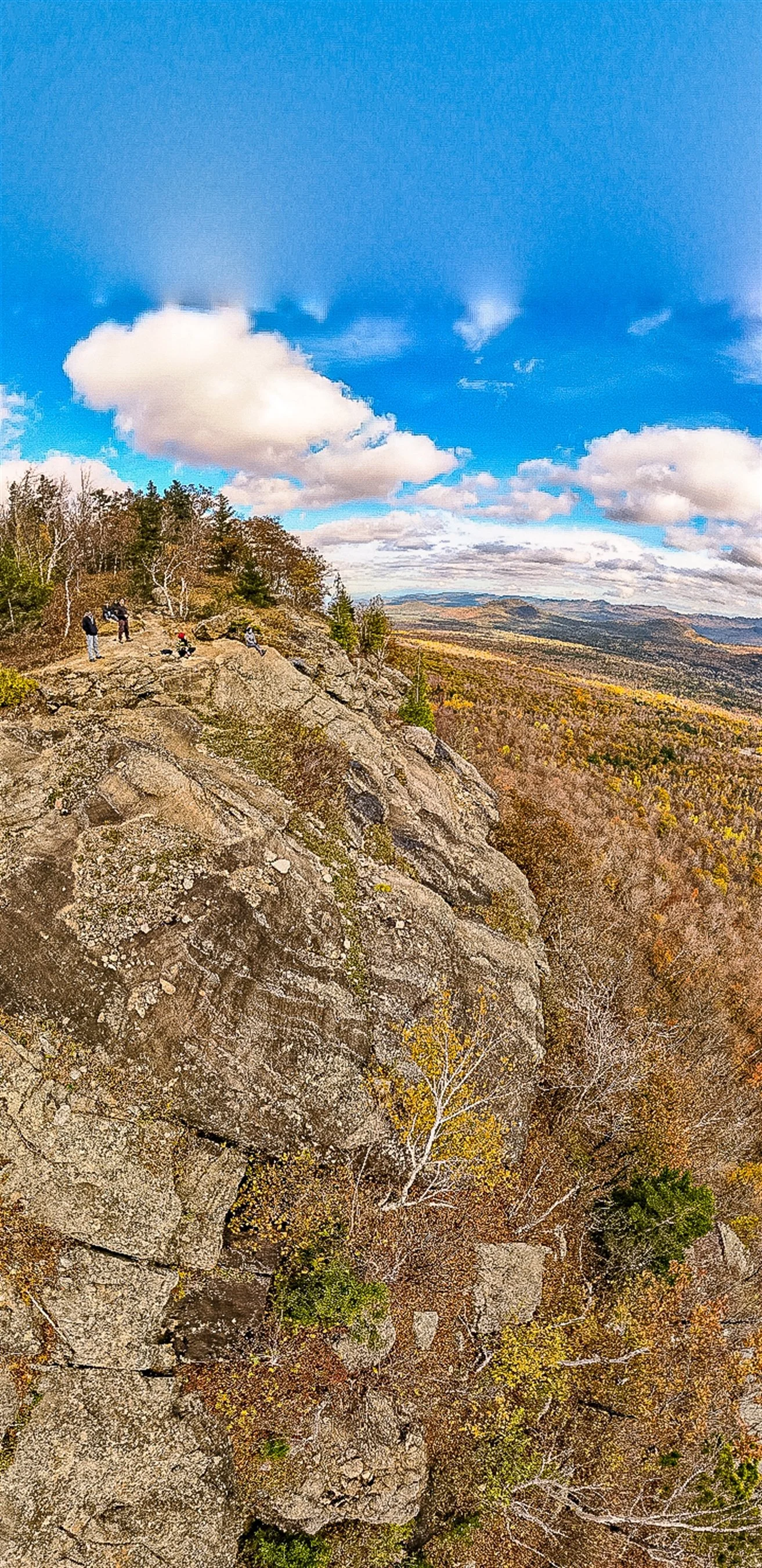 A rocky cliff with hikers on top, overlooking a vast landscape of forests under a partly cloudy sky.