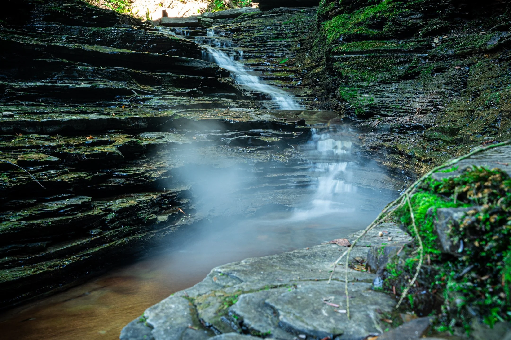 Small waterfall flowing over layered rocks, surrounded by mossy green vegetation in a rocky gorge.