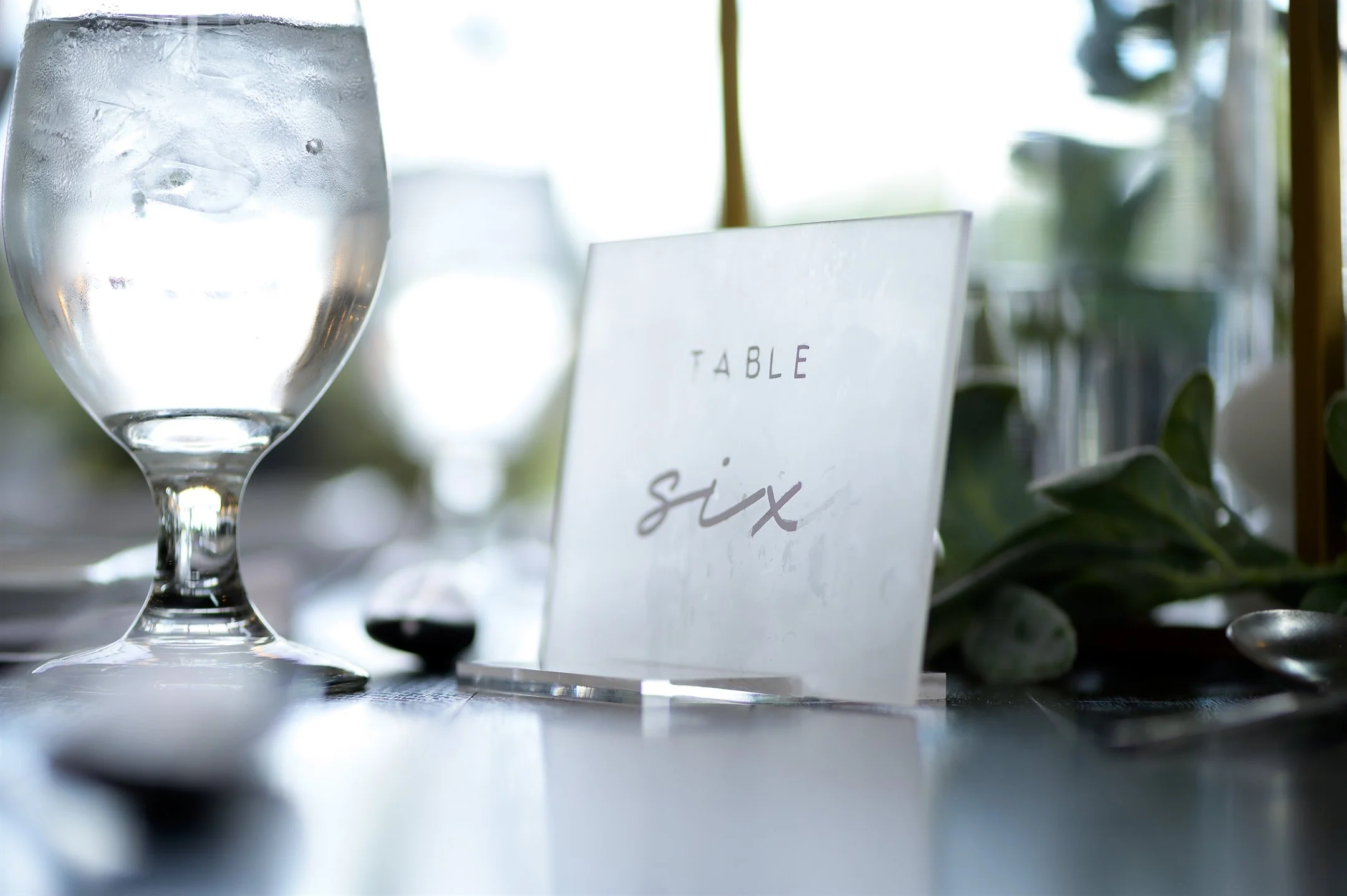 Close-up of a table with a glass of water and a "Table Six" sign, decorated with greenery.