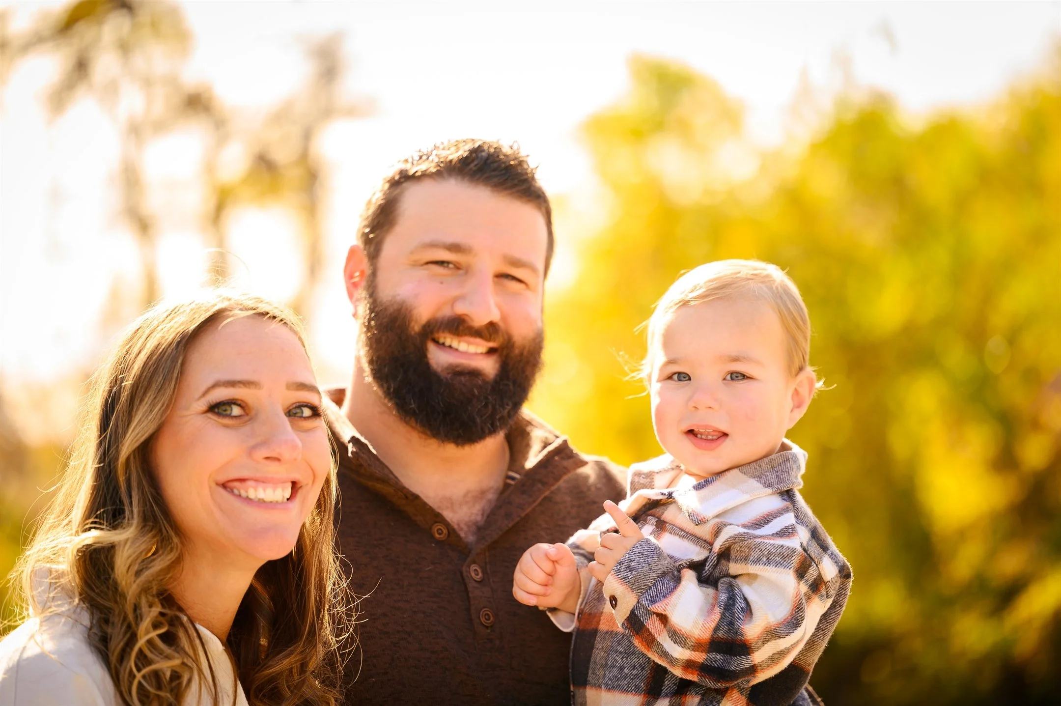 A smiling man, woman, and child outdoors with autumn foliage.