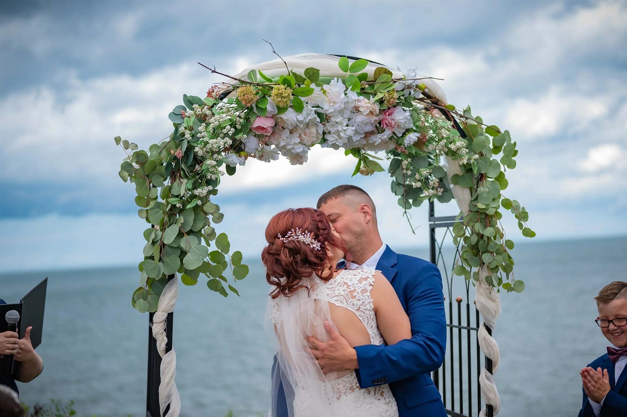 Bride and groom kissing under floral arch at outdoor wedding ceremony, with ocean in background.