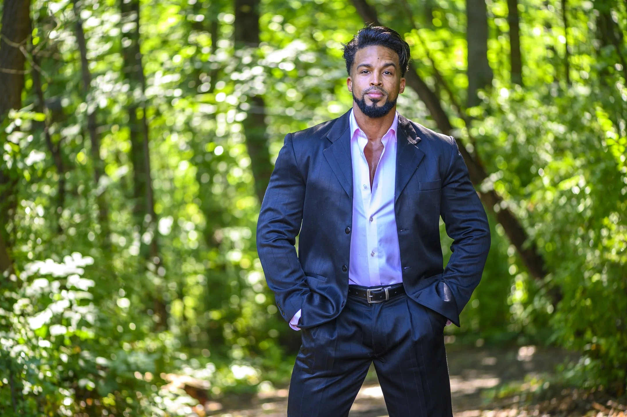 A man in a blue suit standing confidently in a forest path, surrounded by green foliage.