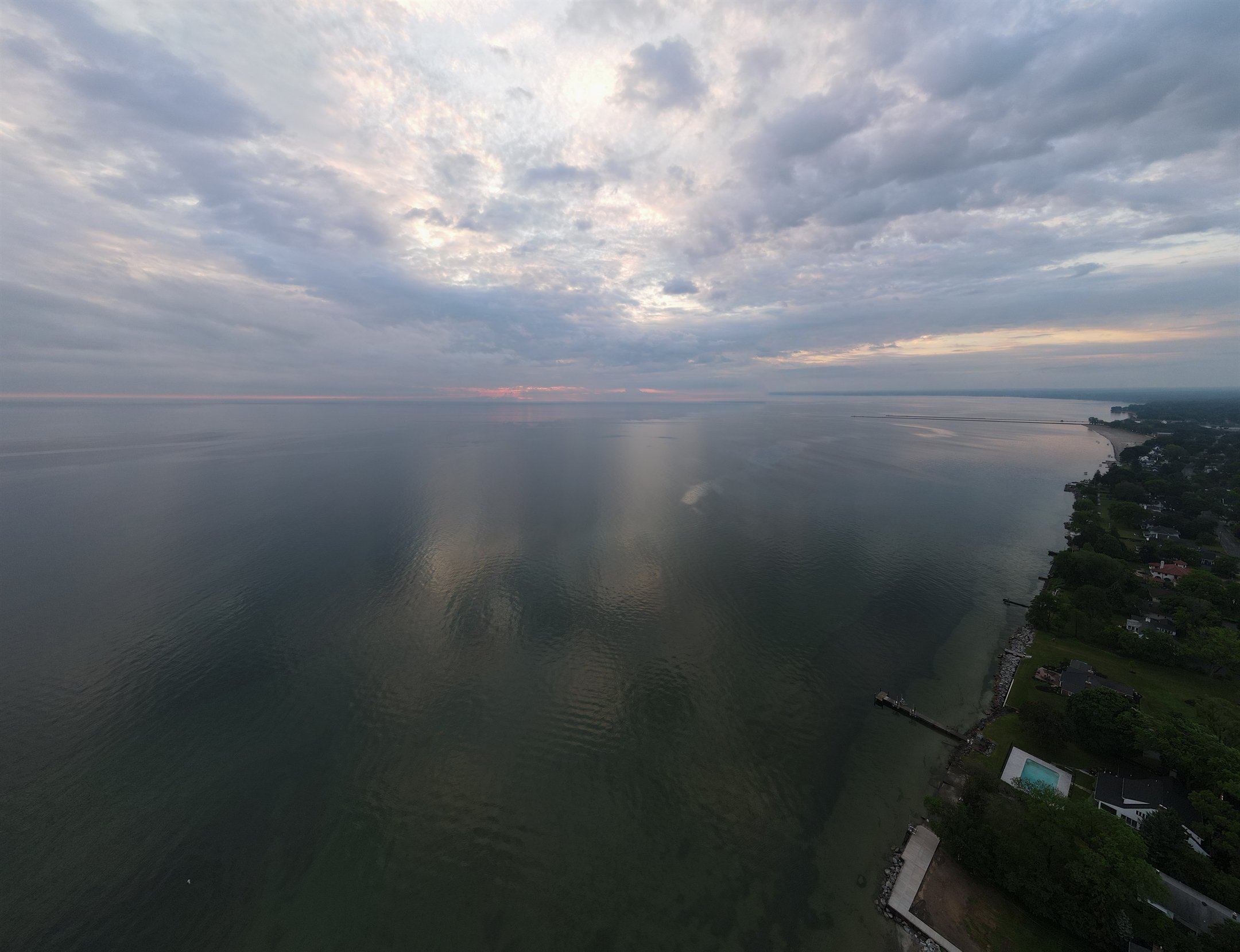 Aerial view of a coastline with calm water and overcast sky, showing faint sunset colors on the horizon. The shoreline is bordered by greenery and residential buildings.
