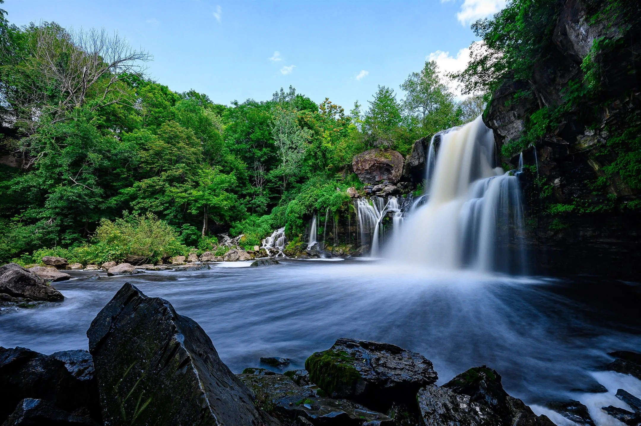 Waterfall flowing into a river surrounded by green trees and rocks on a sunny day.