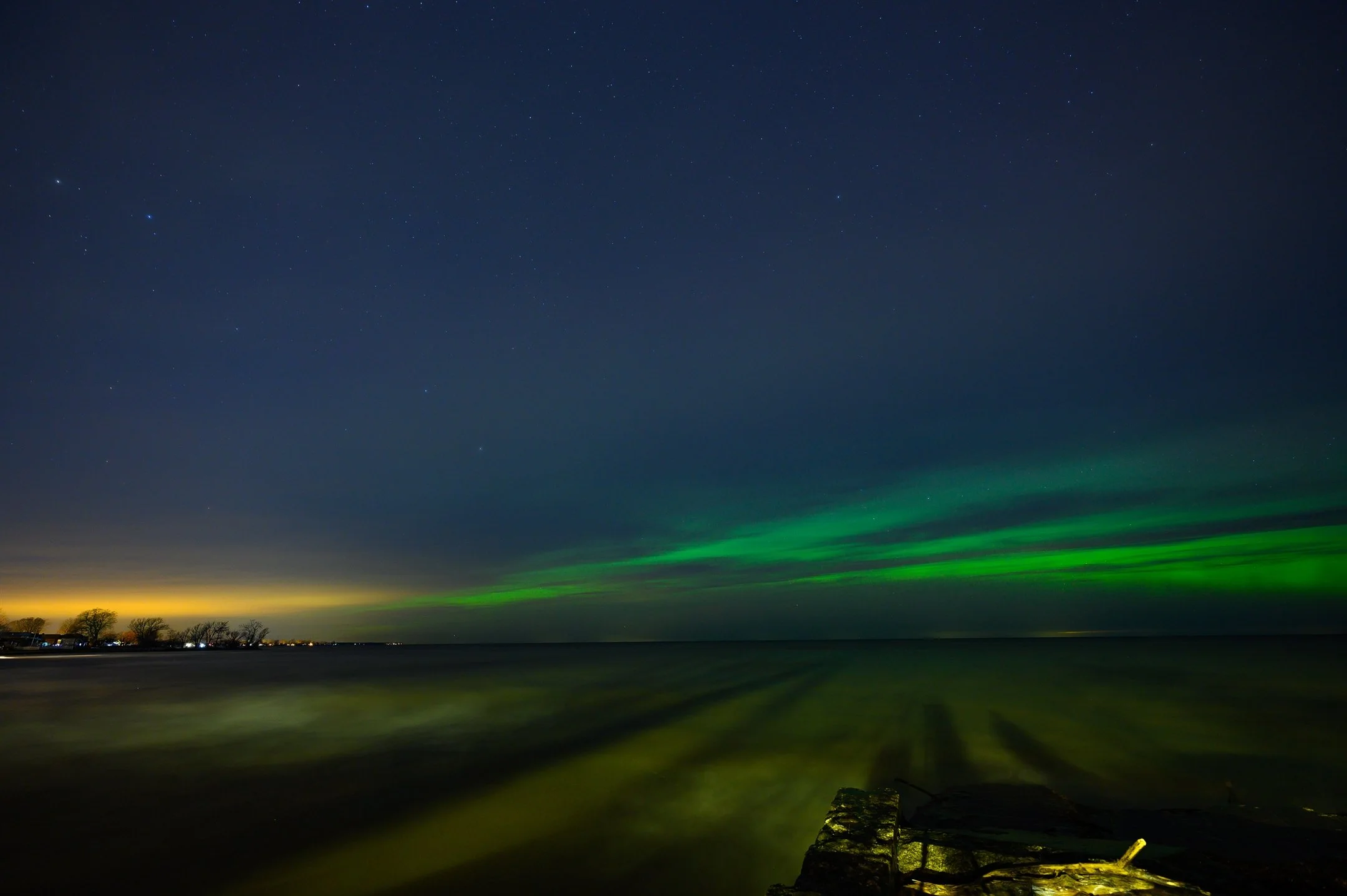 Northern lights over a calm lake with distant trees and stars in the night sky