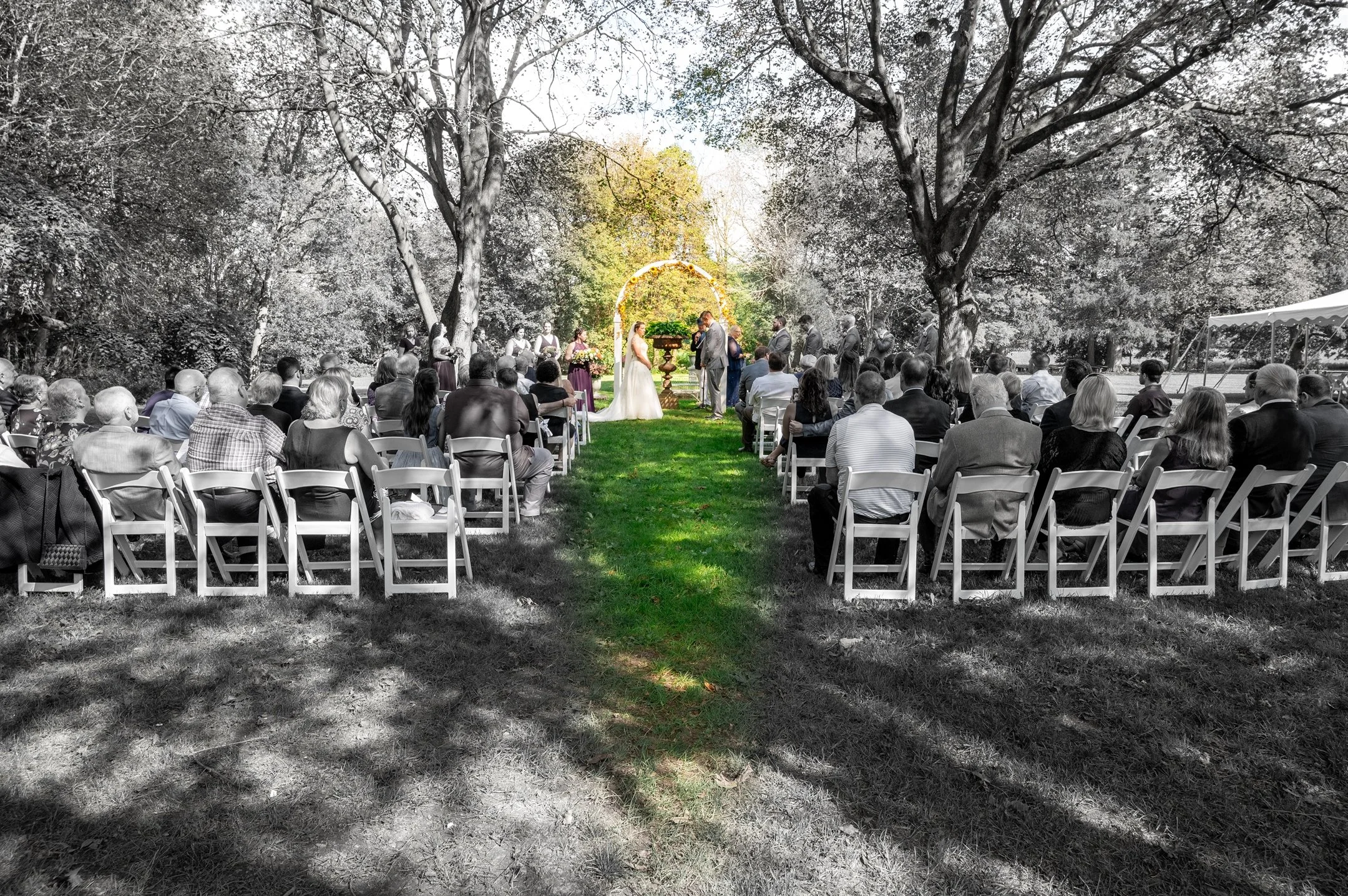 Wedding ceremony outdoors with guests seated on white chairs, bride and groom under a floral arch, surrounded by trees.