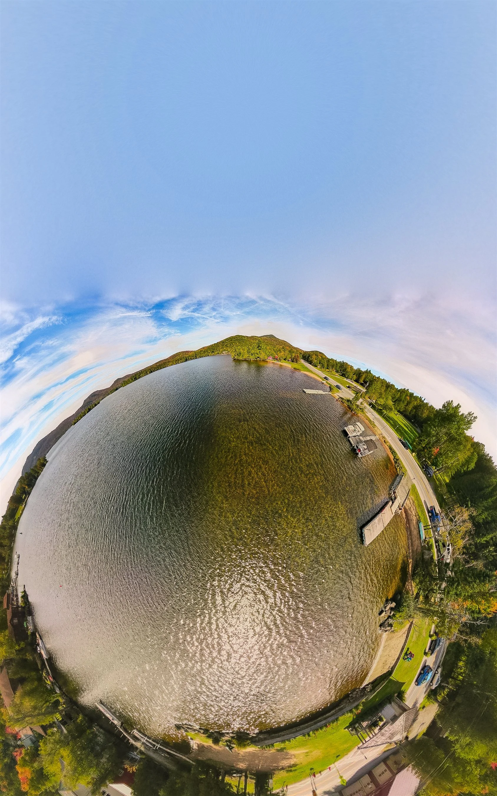 360-degree panoramic view of a lake with surrounding landscape, docks, and trees under a blue sky.