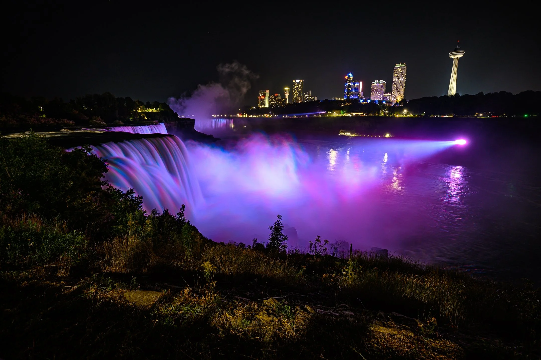 Niagara Falls at night with colorful lights illuminating the waterfalls and a city skyline in the background.