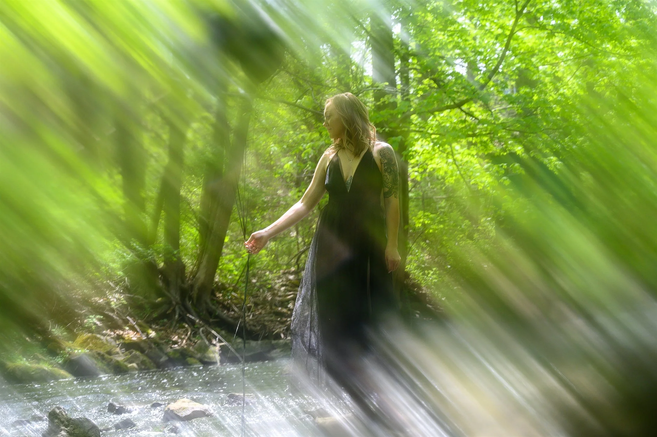 Woman in black dress standing by a stream in a forest with blurred green leaves.