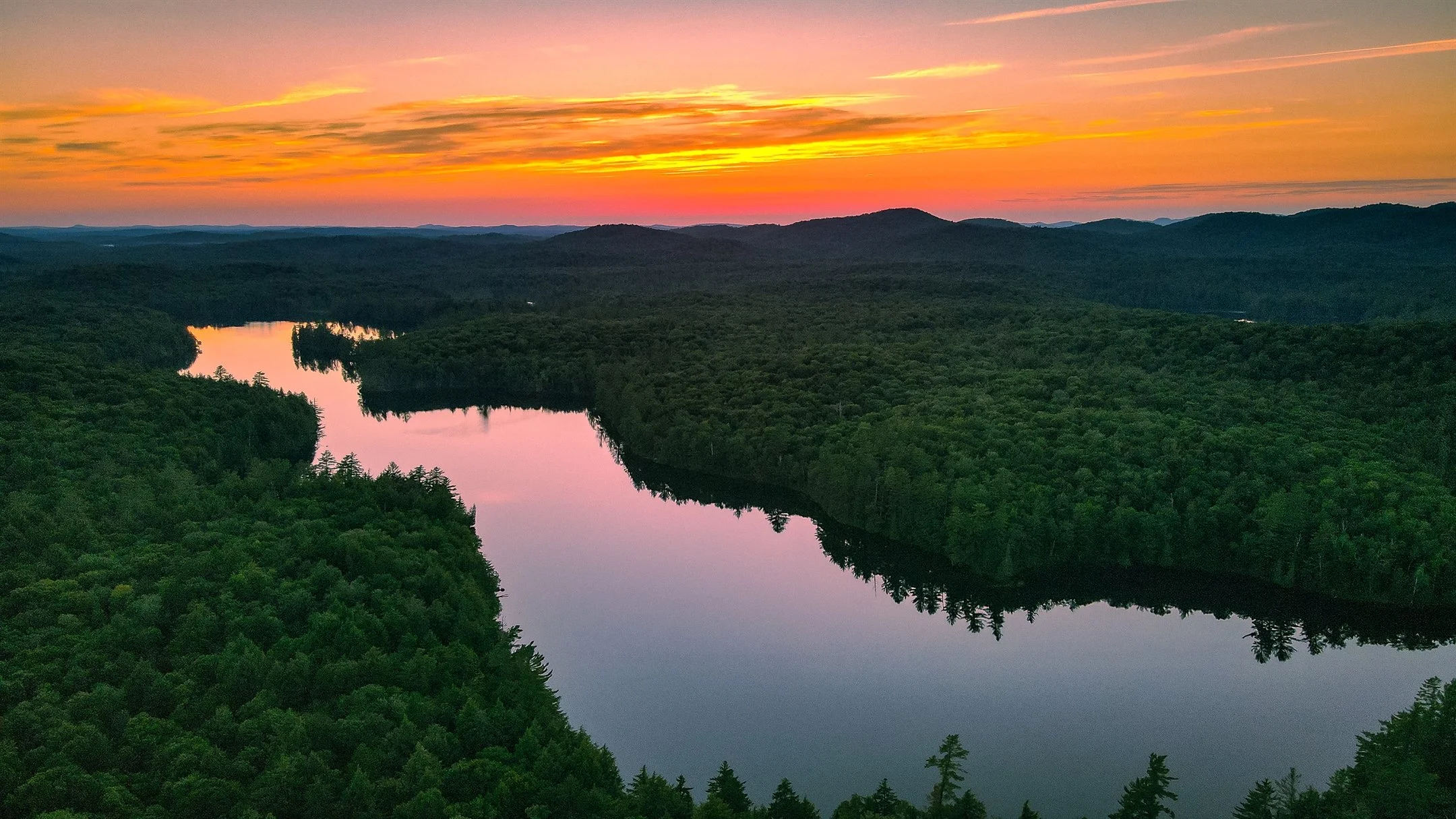 Aerial view of a river winding through a dense forest at sunset, with an orange and pink sky reflecting on the water.