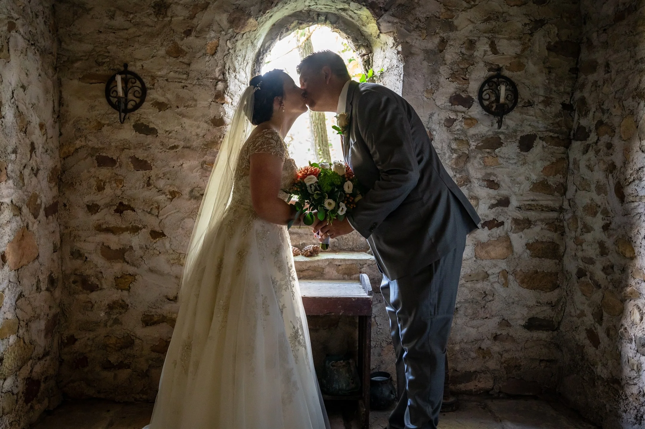 Bride and groom kissing in stone-walled room holding flowers, backlit by a window.
