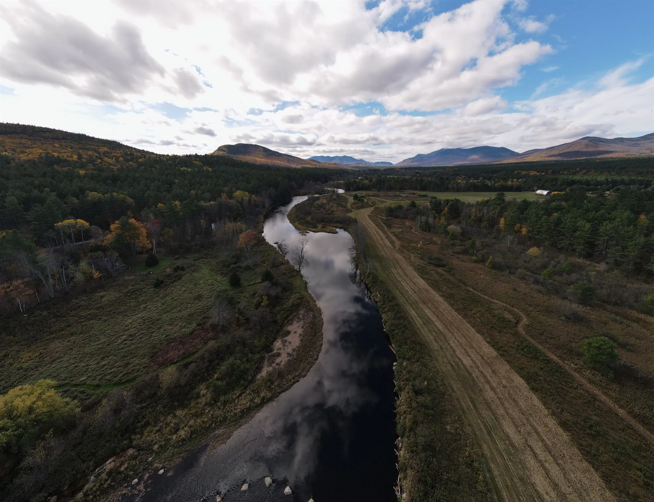 Aerial view of a scenic landscape featuring a winding river, lush green fields, dense forests, and distant mountains under a partly cloudy sky.