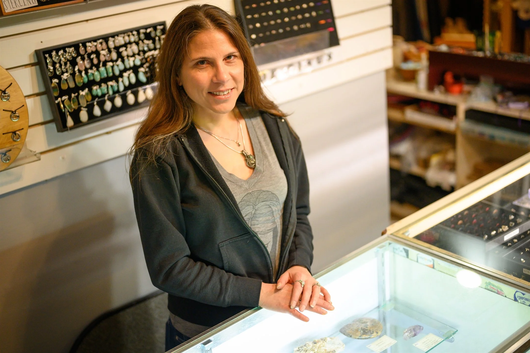 Woman standing behind a glass display case in a jewelry or gem store, with various necklaces, rings, and stones visible.