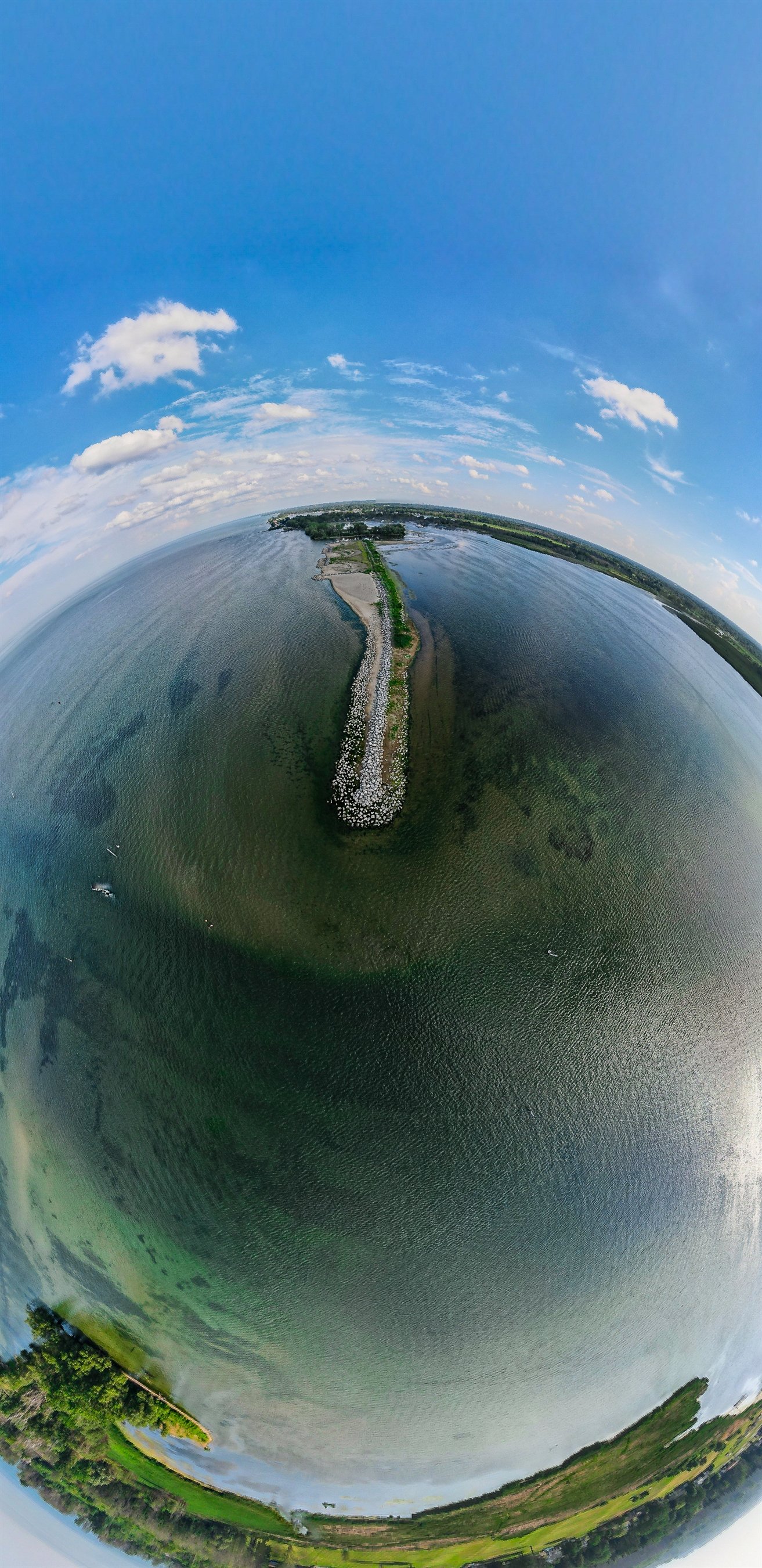 Drone view of a coastal landscape with water, shoreline, and a long stone causeway extending into the ocean, surrounded by greenery and clouds in a blue sky.