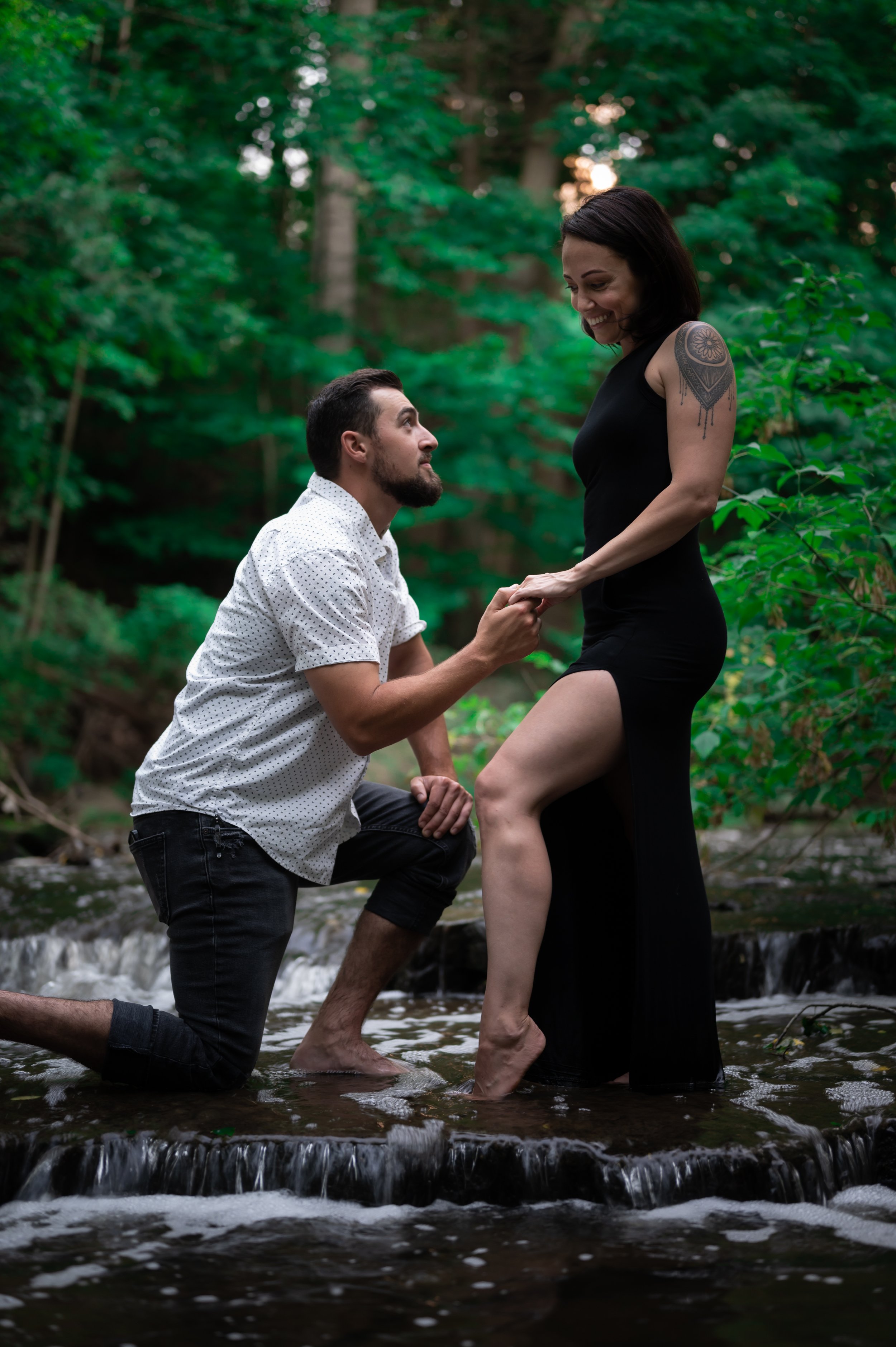 Man kneeling proposing to woman in black dress by a stream in a forest.
