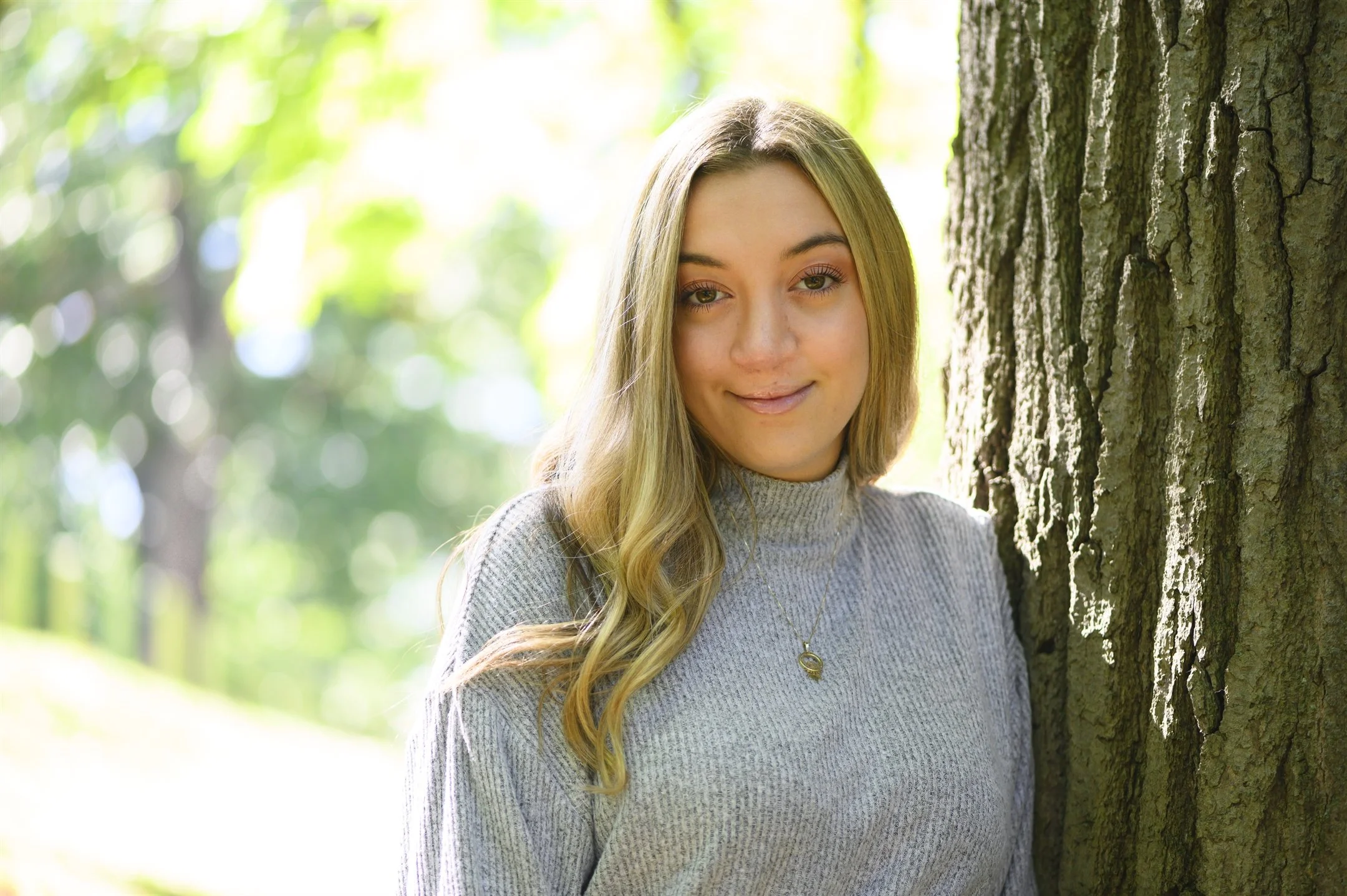 A woman with long blonde hair wearing a gray sweater, standing next to a tree in a sunny outdoor setting.